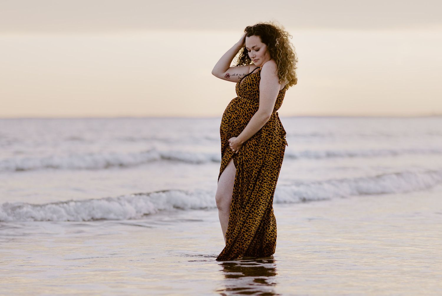 Artistic series of beach maternity photos with subject walking in animal print dress at sunset.