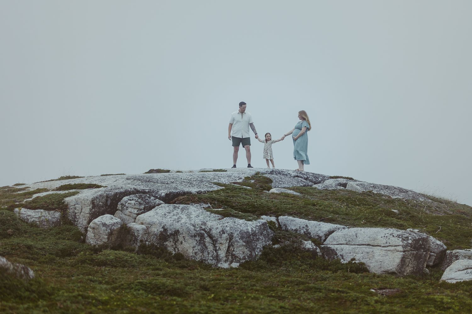 Family of three holding hands on rocky hilltop in misty landscape.