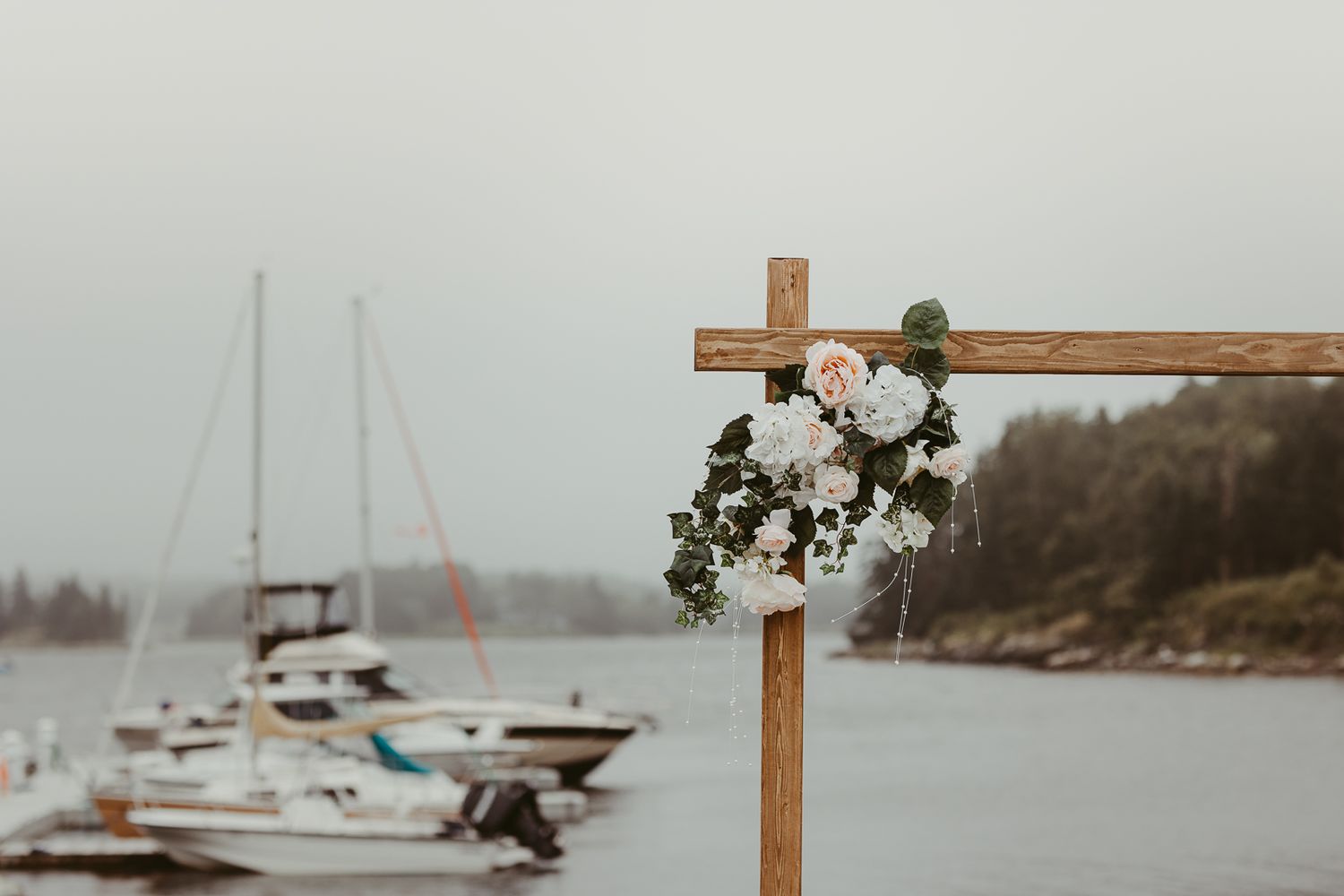 A wooden cross decorated with flowers stands against a marina backdrop with boats in the water.