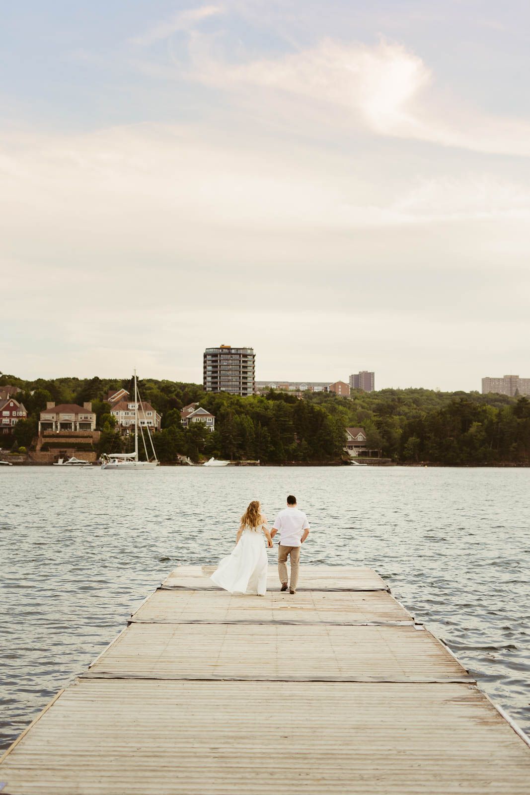 Newlyweds walk along a wooden pier with waterfront homes and boats visible in the background.