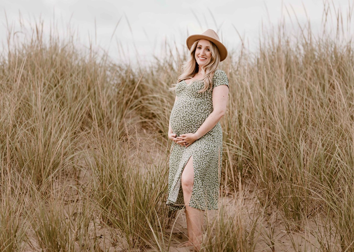 Pregnant woman in floral dress and straw hat posing among tall beach grass during maternity photoshoot.