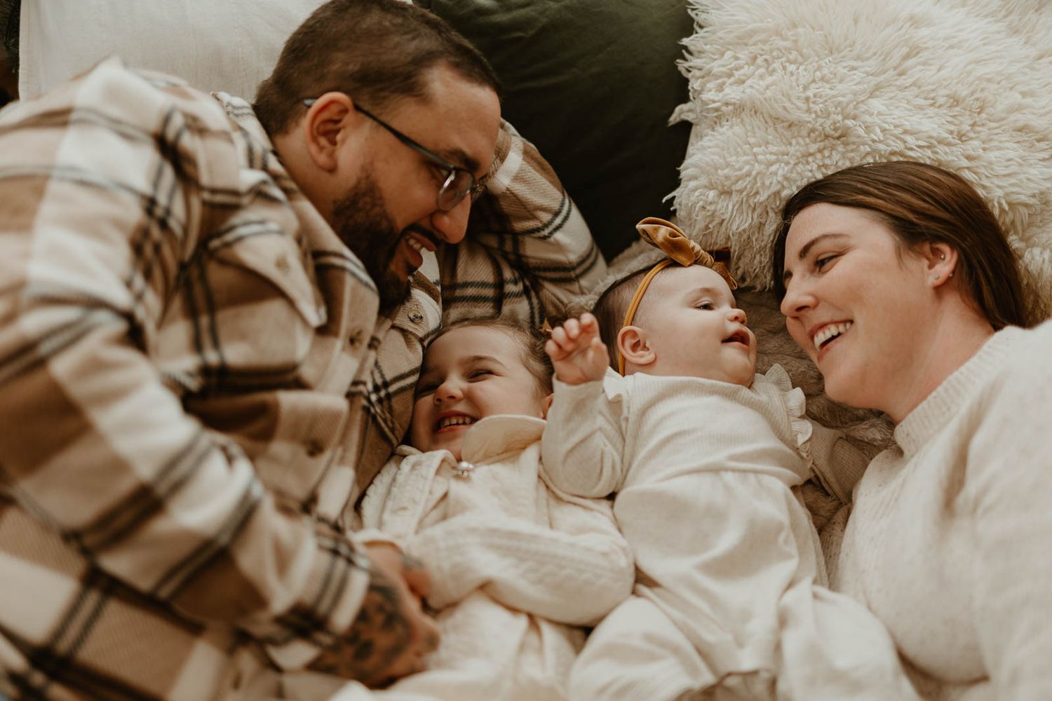 Happy family with two babies lying together on a cozy bed, smiling and laughing in matching white outfits.