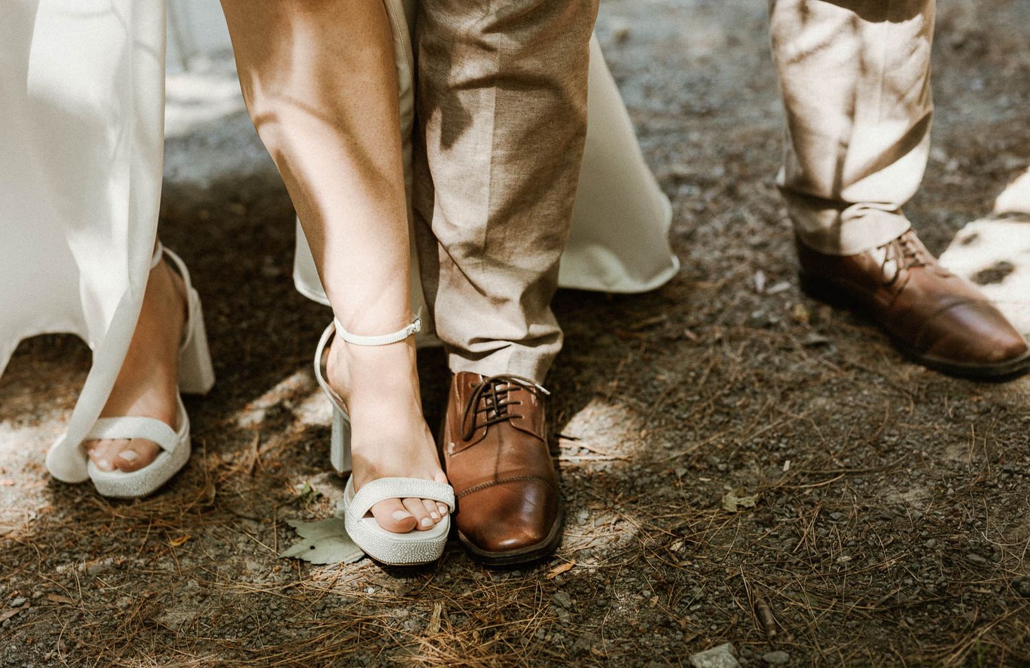 Close-up shot of bride and groom's contrasting footwear with white peep-toe heels and brown leather dress shoes on dirt ground.