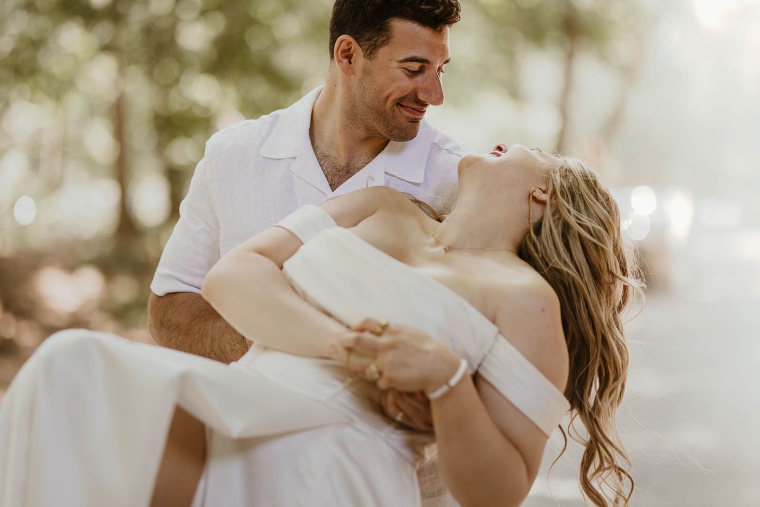 A groom in white carries his bride outdoors, both smiling joyfully in a romantic wedding photo.