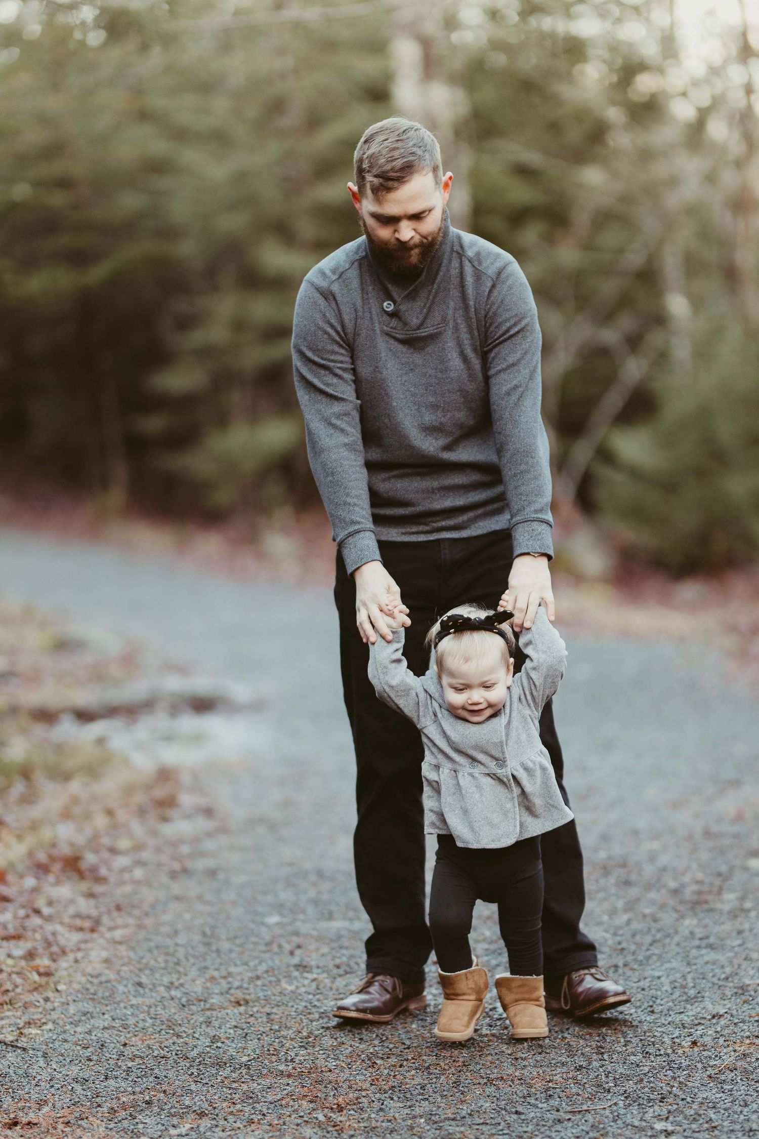 Parent helping small child learn to walk on a gravel path while wearing matching grey sweaters.