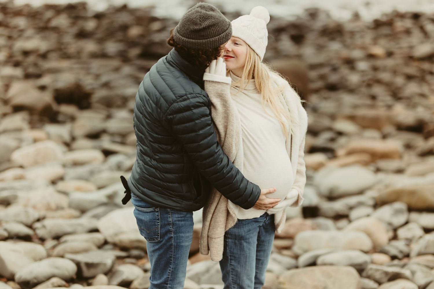 A couple shares an intimate moment on a rocky beach during a winter maternity photoshoot.