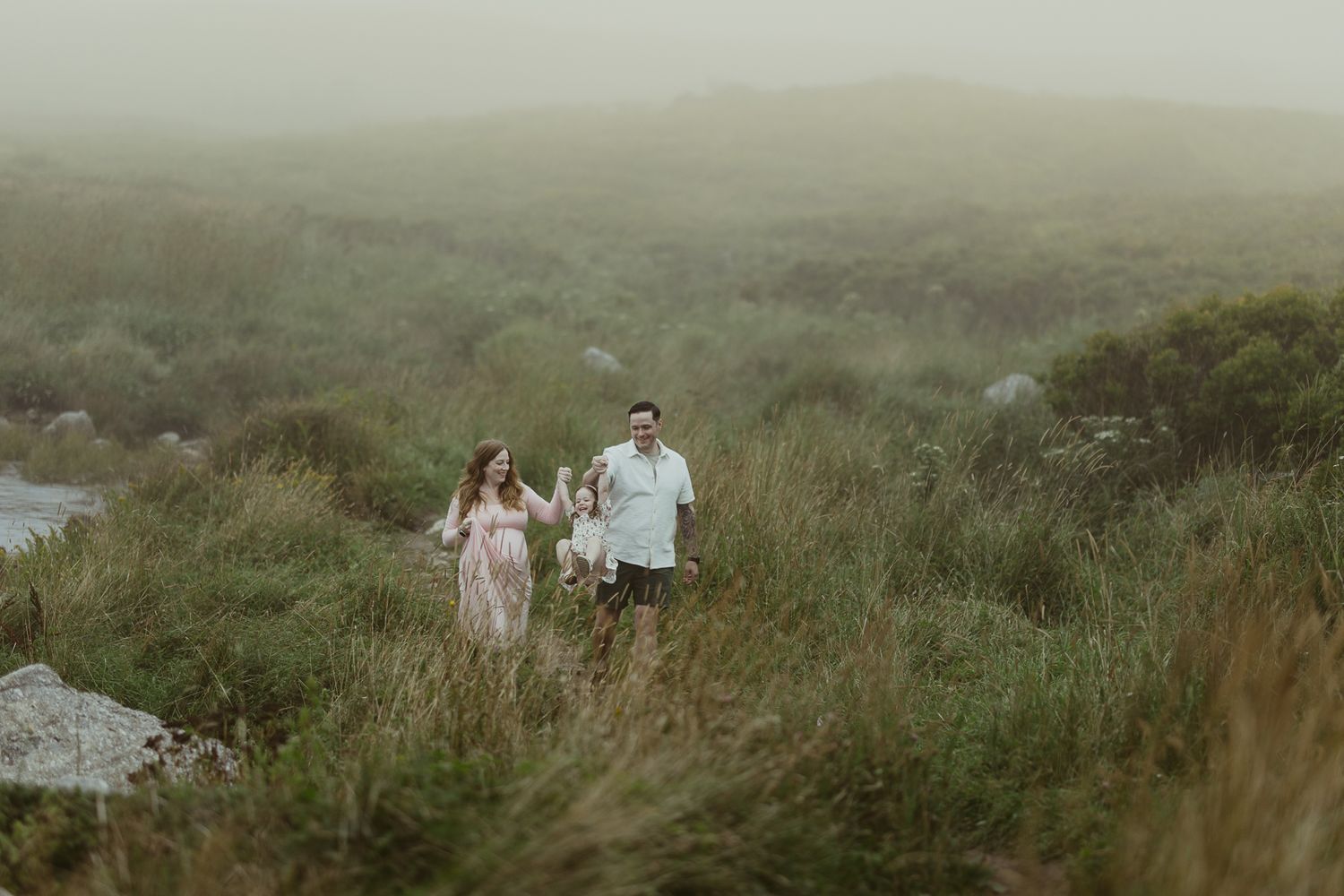 A family walks through misty green hills, with a woman in a pink dress and a man in a light shirt carrying a child.