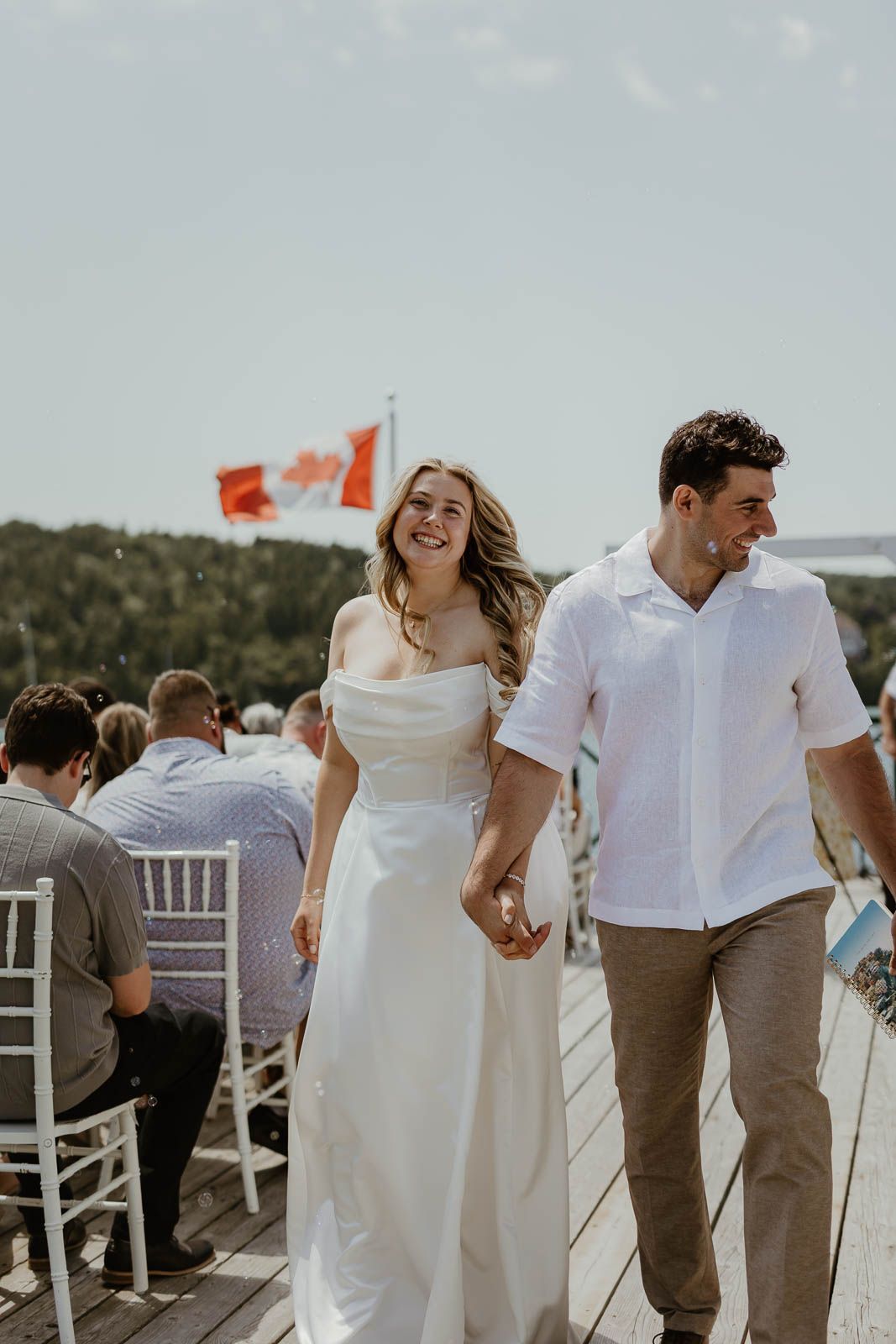 A couple walks hand in hand during their outdoor wedding ceremony with guests and Canadian flags in the background.