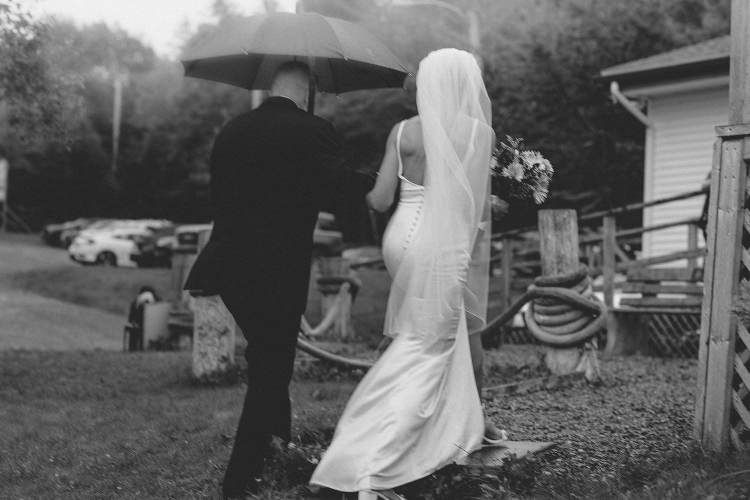Black and white image of couple walking with umbrella during wedding celebration.