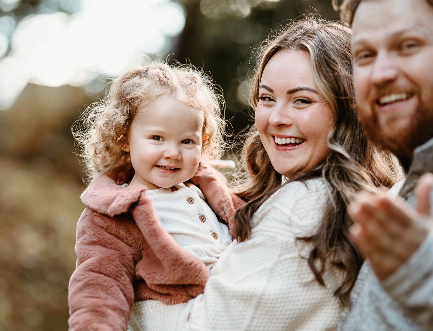 Happy family portrait with smiling mother holding curly-haired toddler girl in pink jacket outdoors.