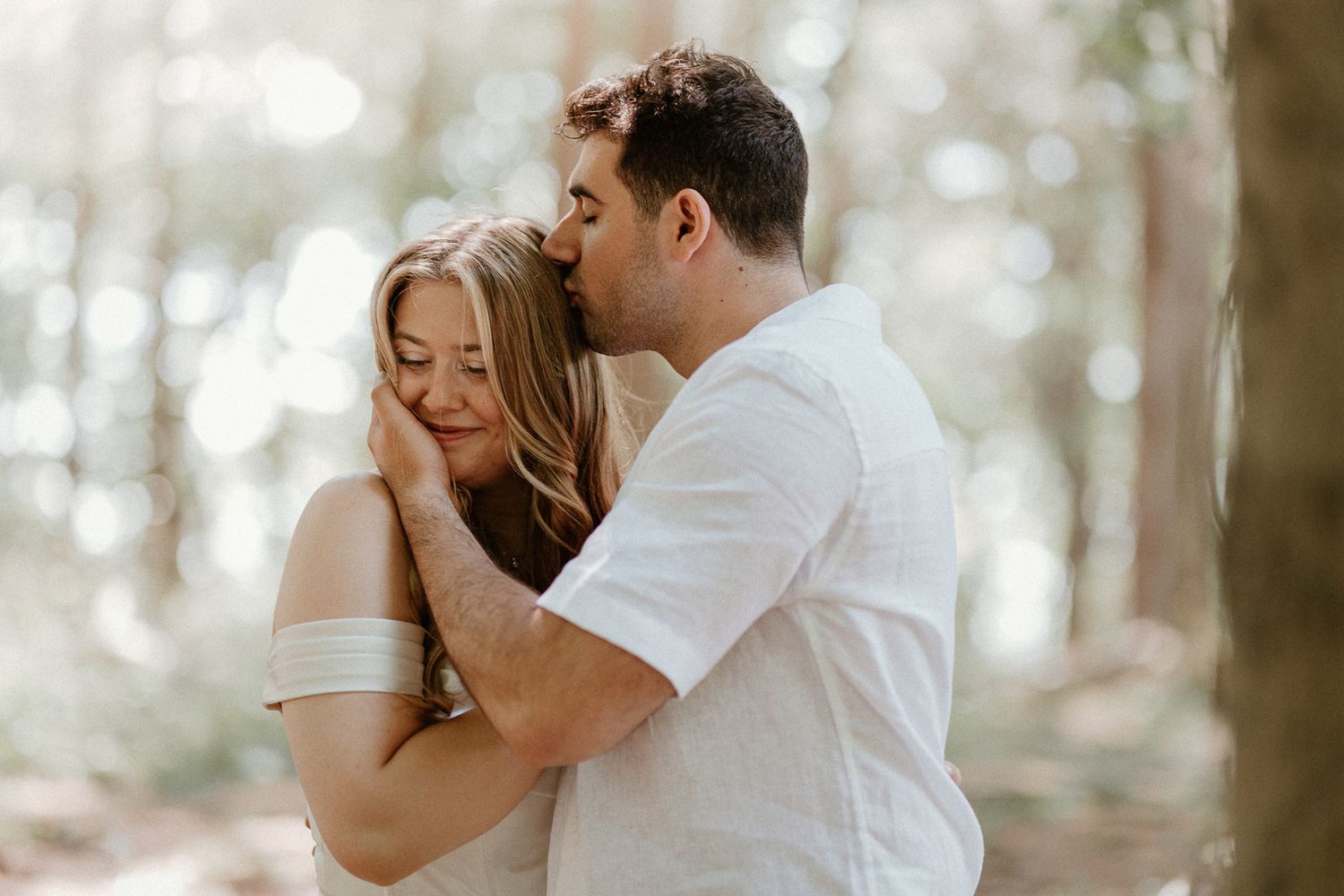 Couple sharing an intimate embrace during engagement photoshoot in sunlit forest.