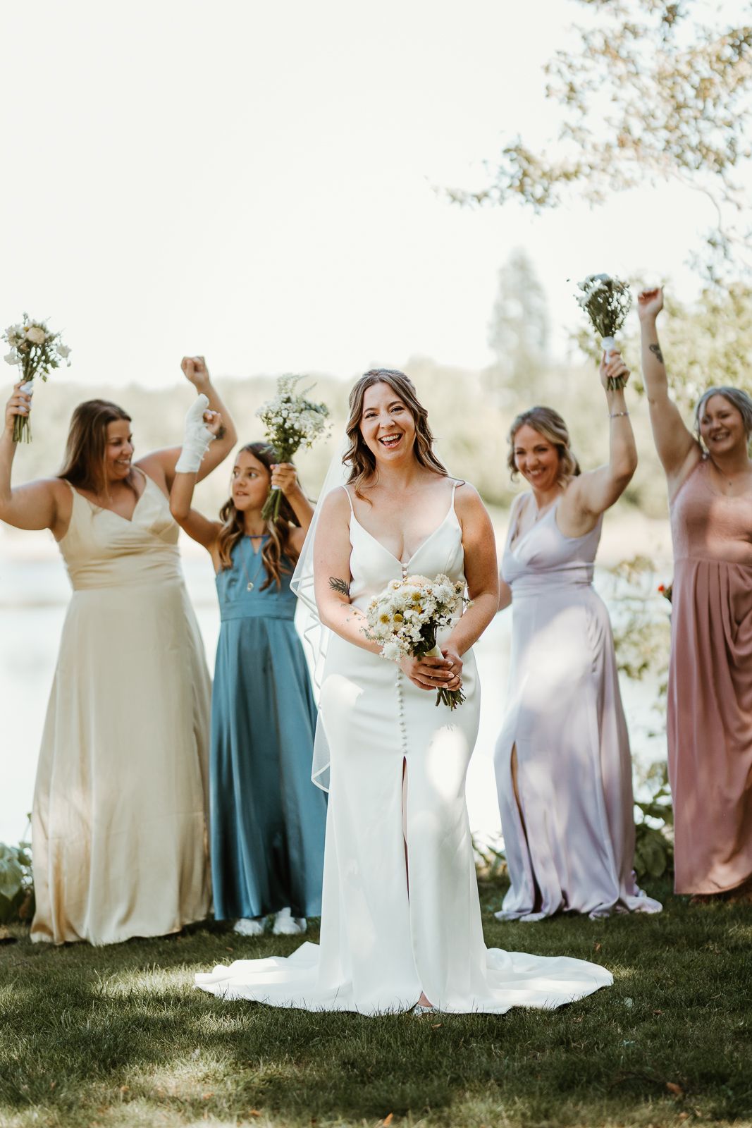 Group of bridesmaids in pastel dresses celebrating joyfully on grassy lawn during outdoor wedding.