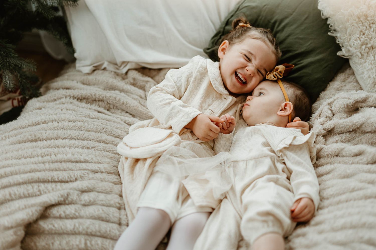 Two adorable baby girls in white outfits laughing together on a cozy blanket, sharing a joyful moment.