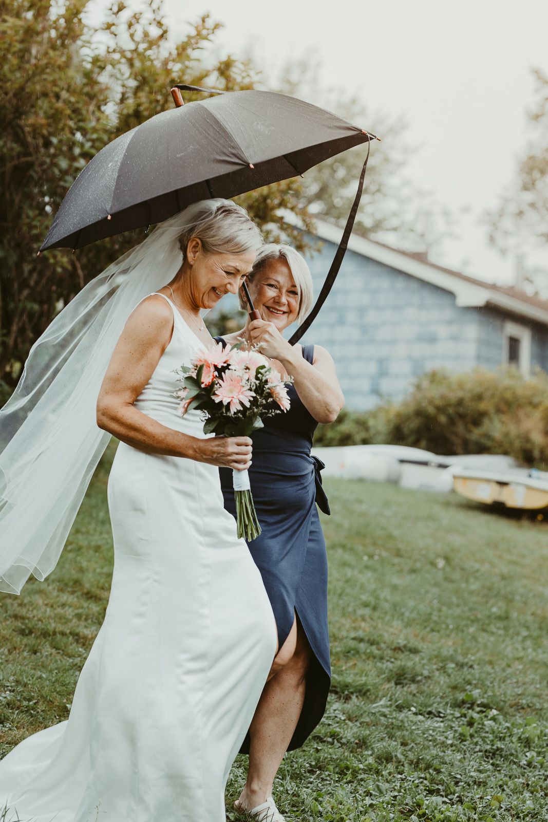 Two women share a joyful moment under a black umbrella, one in a wedding dress holding a bouquet.
