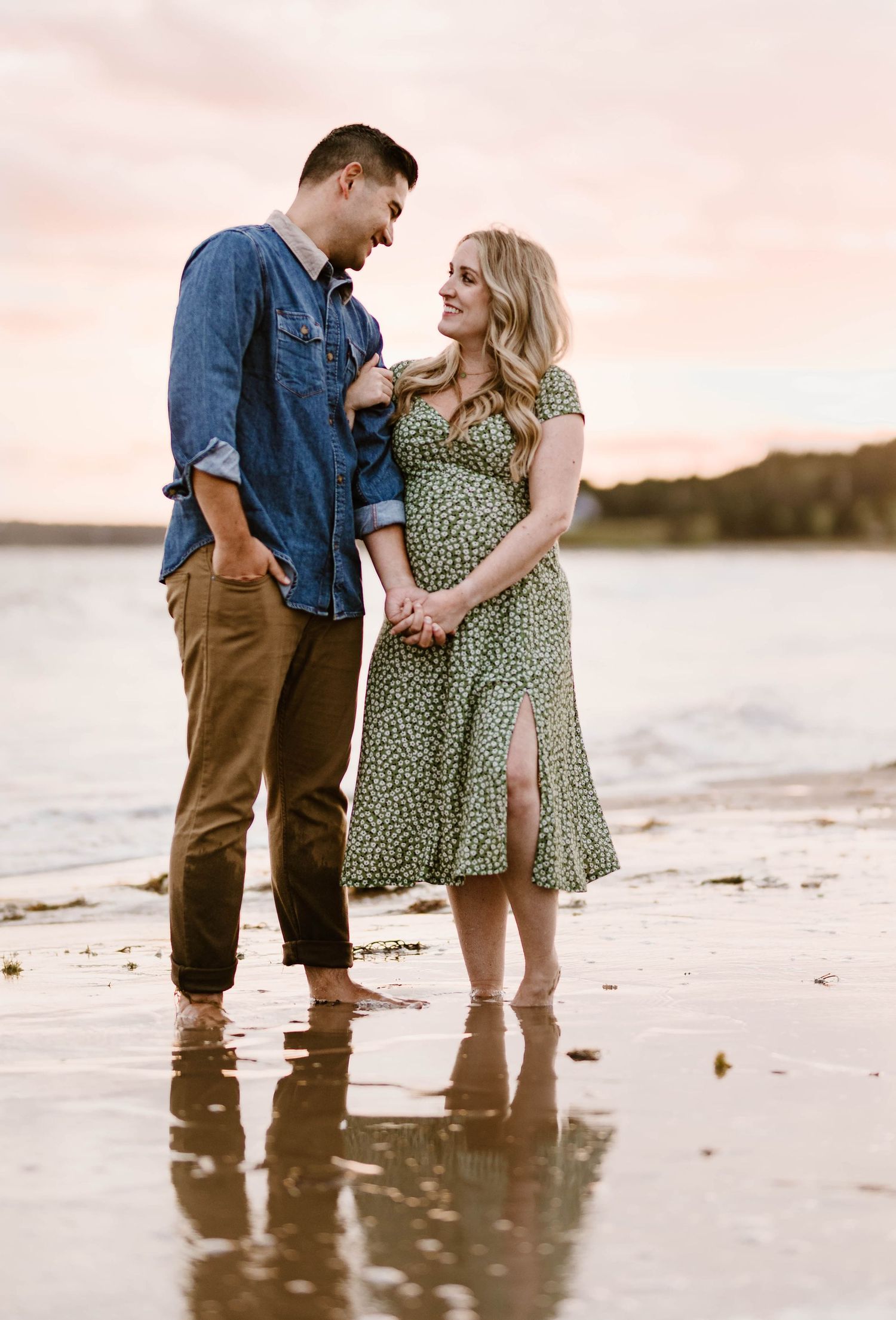Beach maternity session features couple standing in shallow water at sunset.
