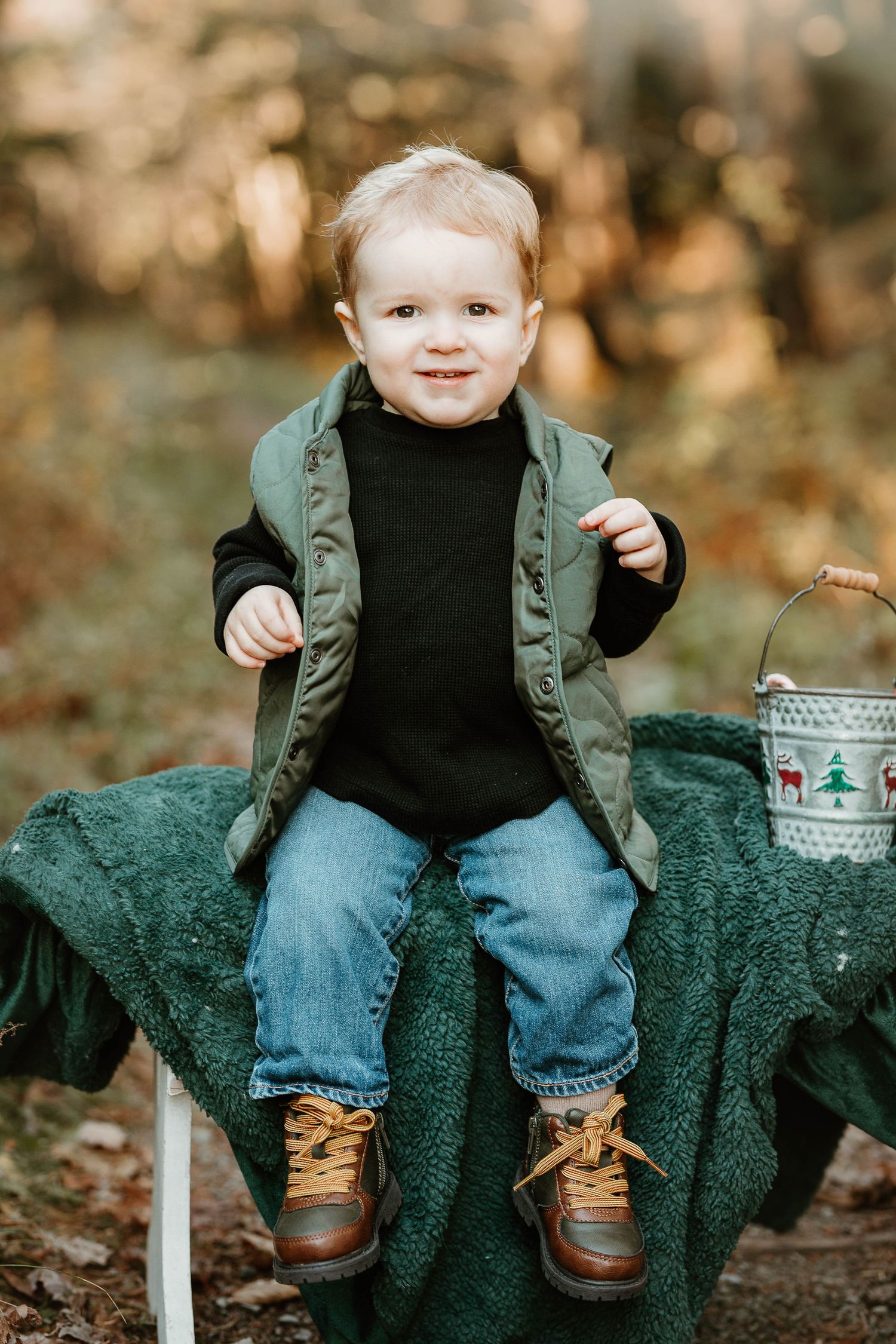 Smiling toddler boy in green vest, black turtleneck, jeans, and brown boots sits outdoors on green blanket with holiday pail.