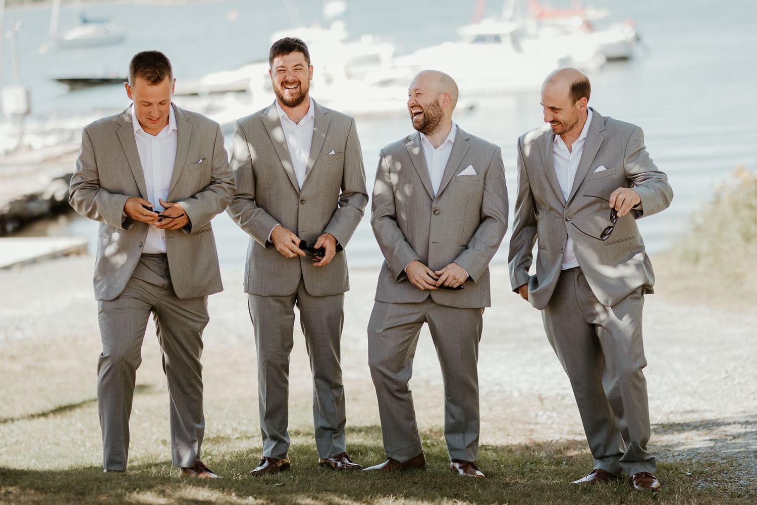 Four groomsmen in matching gray suits share a candid laughing moment by the waterfront.