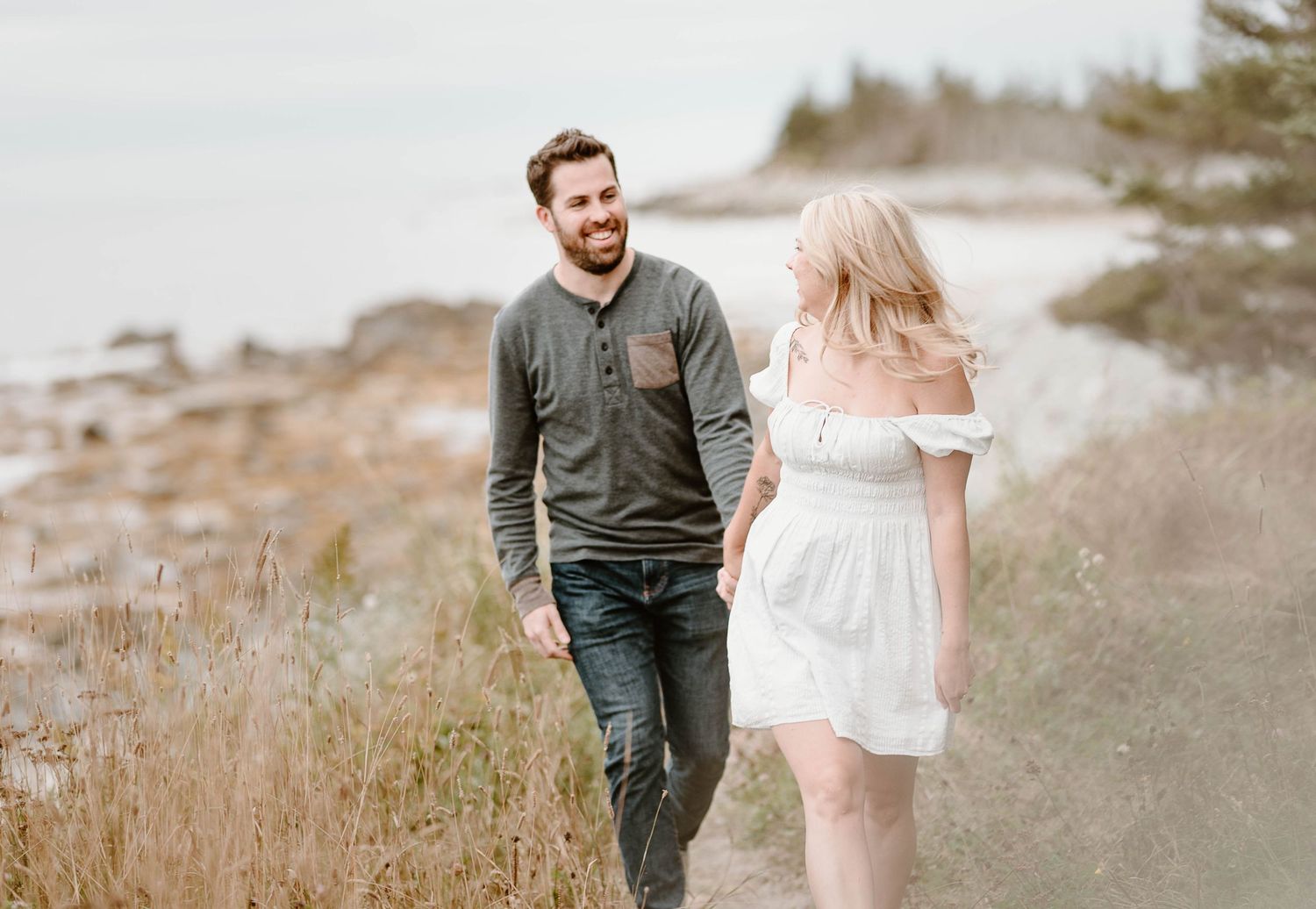 Couple walking on a scenic beach path, man smiling at blonde woman in white dress, coastal landscape background.