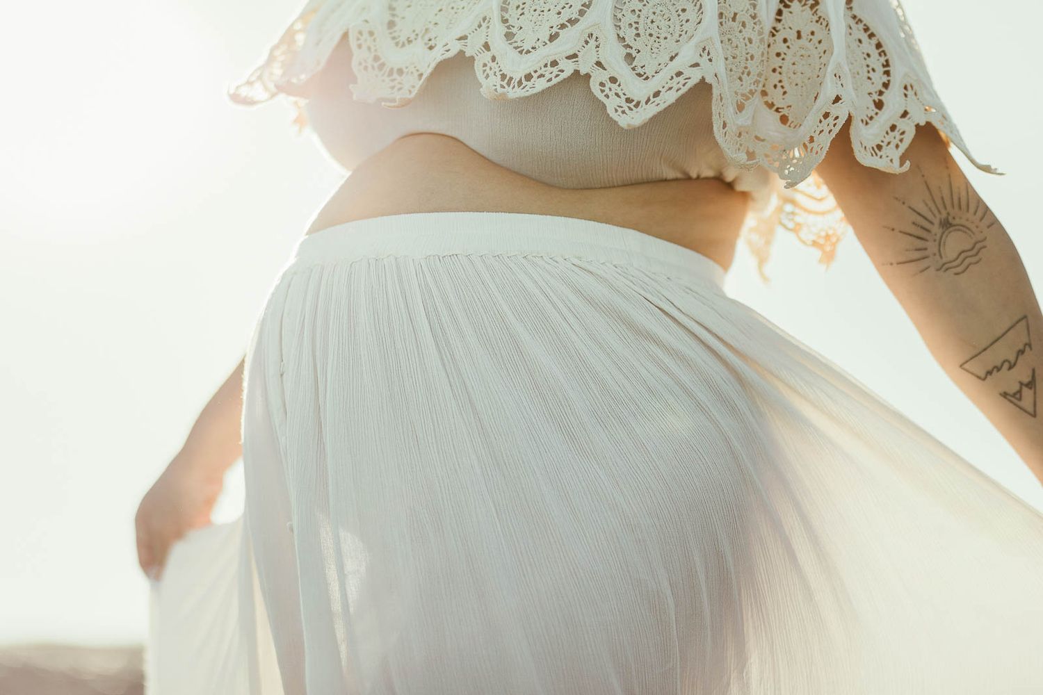 A pregnant mother in a white lace dress stands against soft natural light in a romantic maternity portrait.