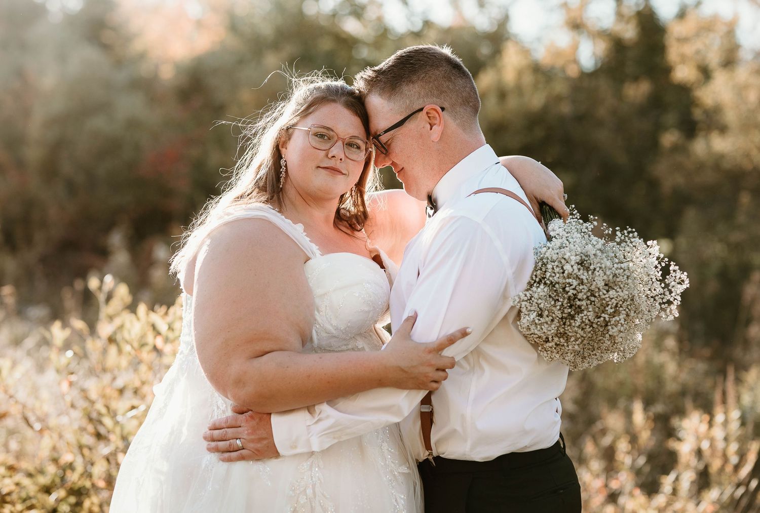 Couple in wedding attire embracing outdoors, bride in white gown holding baby's breath bouquet, groom in white shirt.