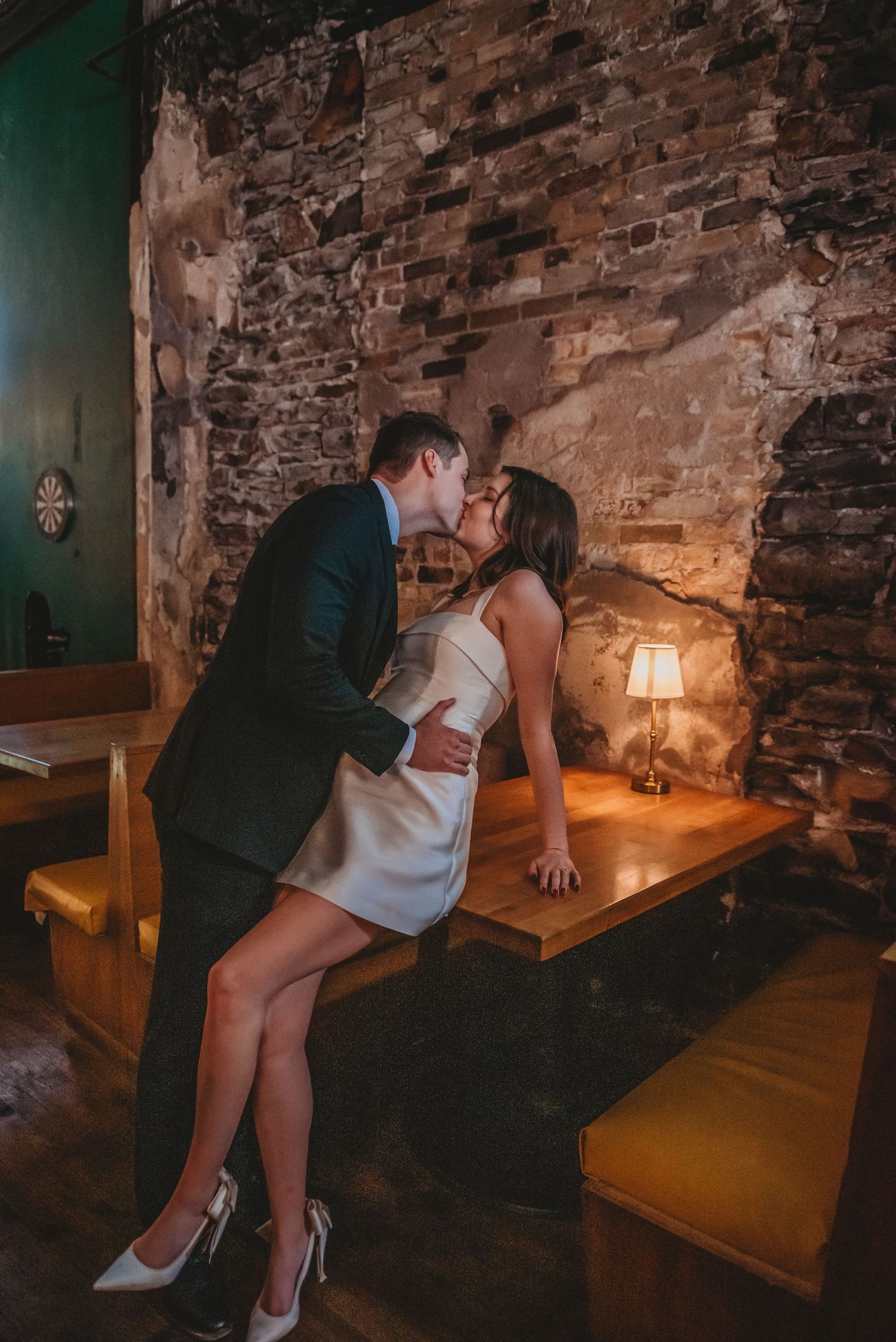 A romantic couple shares an intimate moment in a dimly lit rustic restaurant with exposed brick walls and wooden seating.