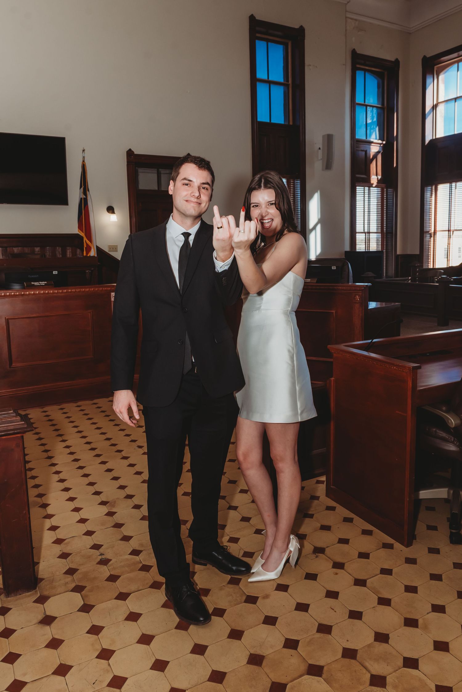 A couple poses together in a courthouse hallway with checkered tile floors and large windows.