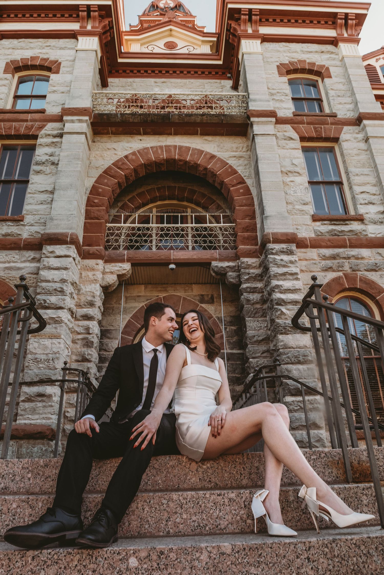 A couple sits on stone steps of a historic Victorian-style building with red brick and stone architectural details.
