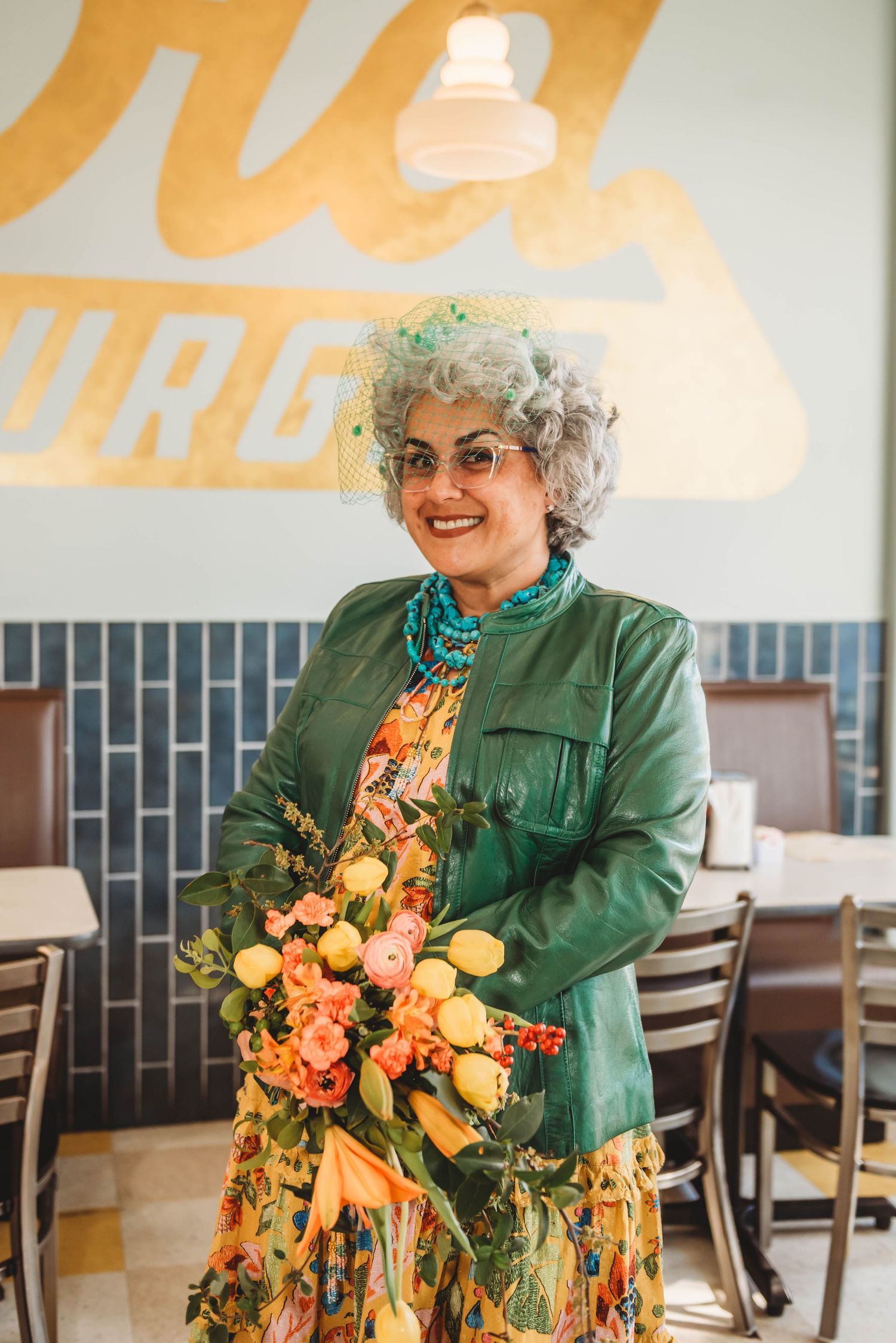 A person in a green jacket and glasses holds a vibrant orange and yellow floral arrangement in a bright interior space.
