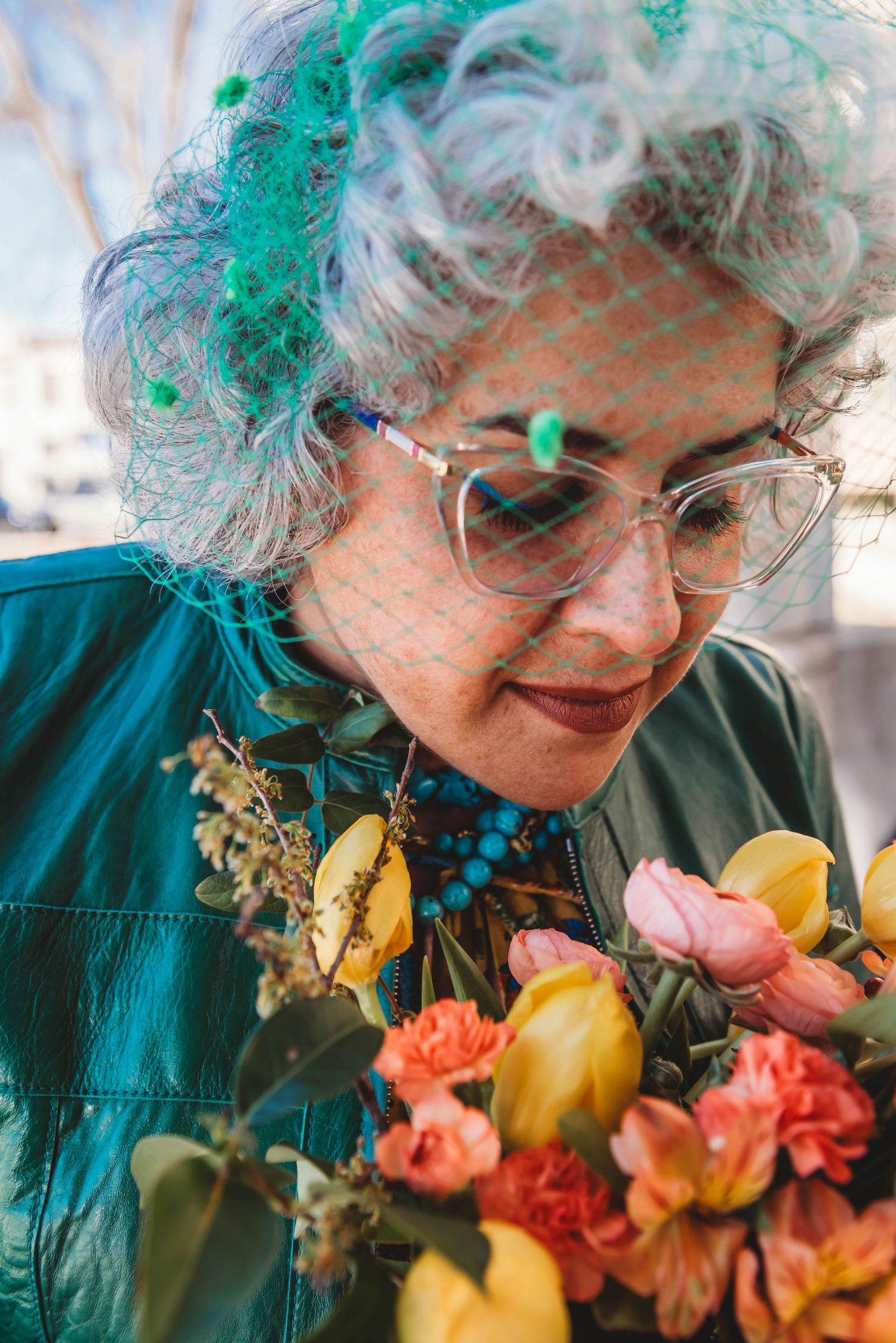 A person with teal-tinted hair and glasses admires a vibrant bouquet of orange and yellow flowers while wearing a green jacket.