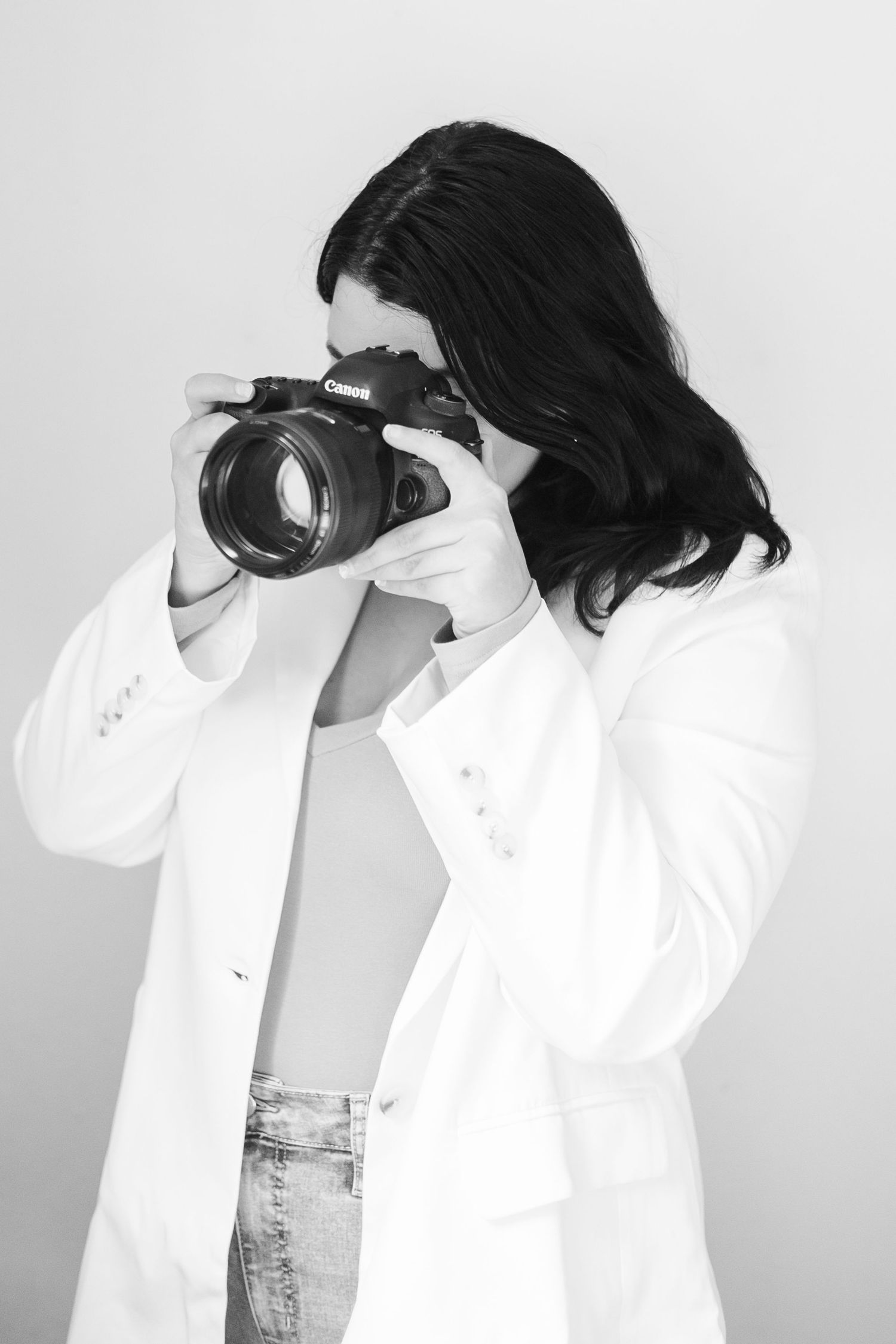 A photographer wearing a white blazer and jeans takes a photo with a professional camera against a gray background.