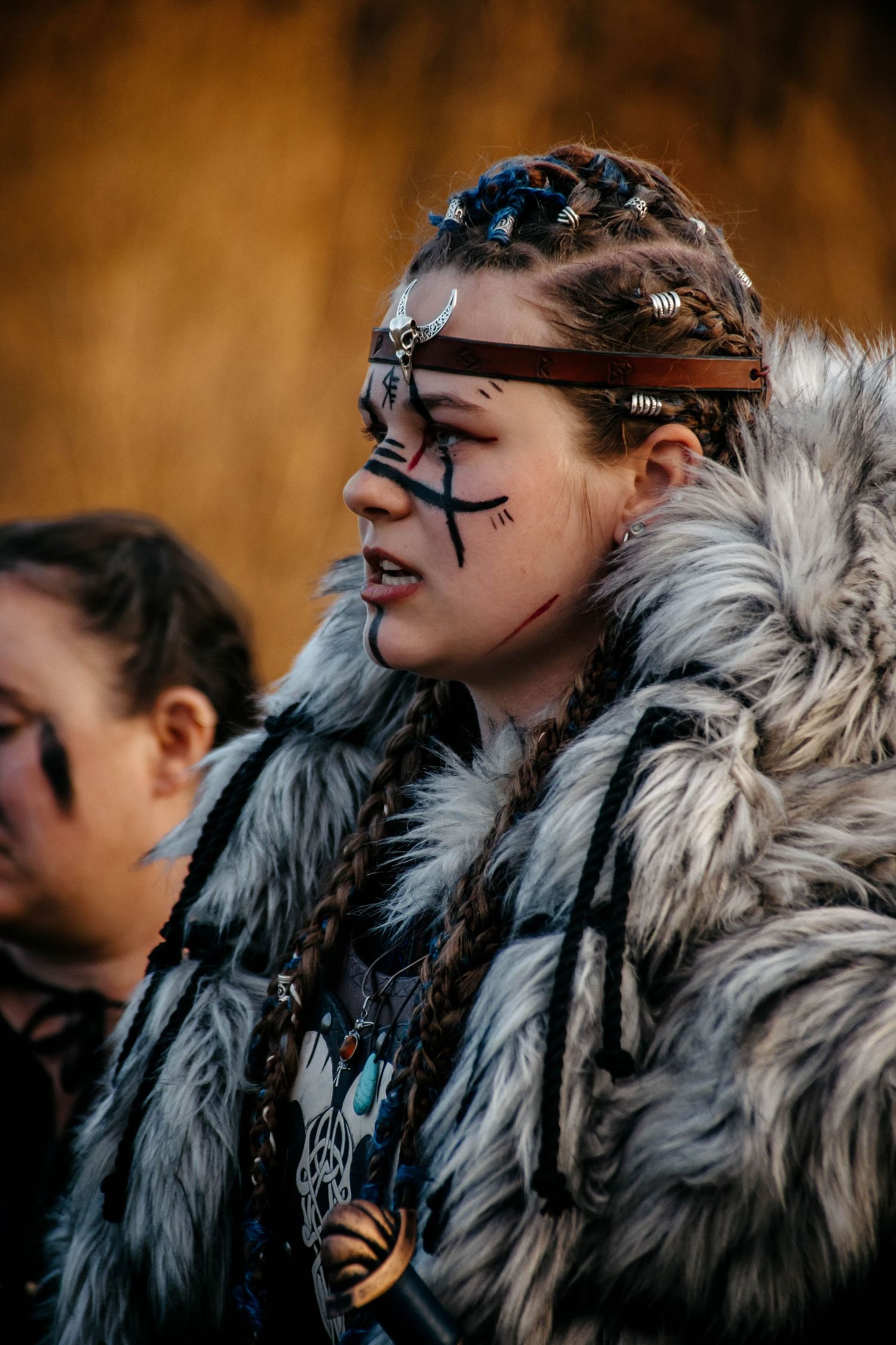 Person wearing tribal face paint and fur cloak poses against blurred autumn background.