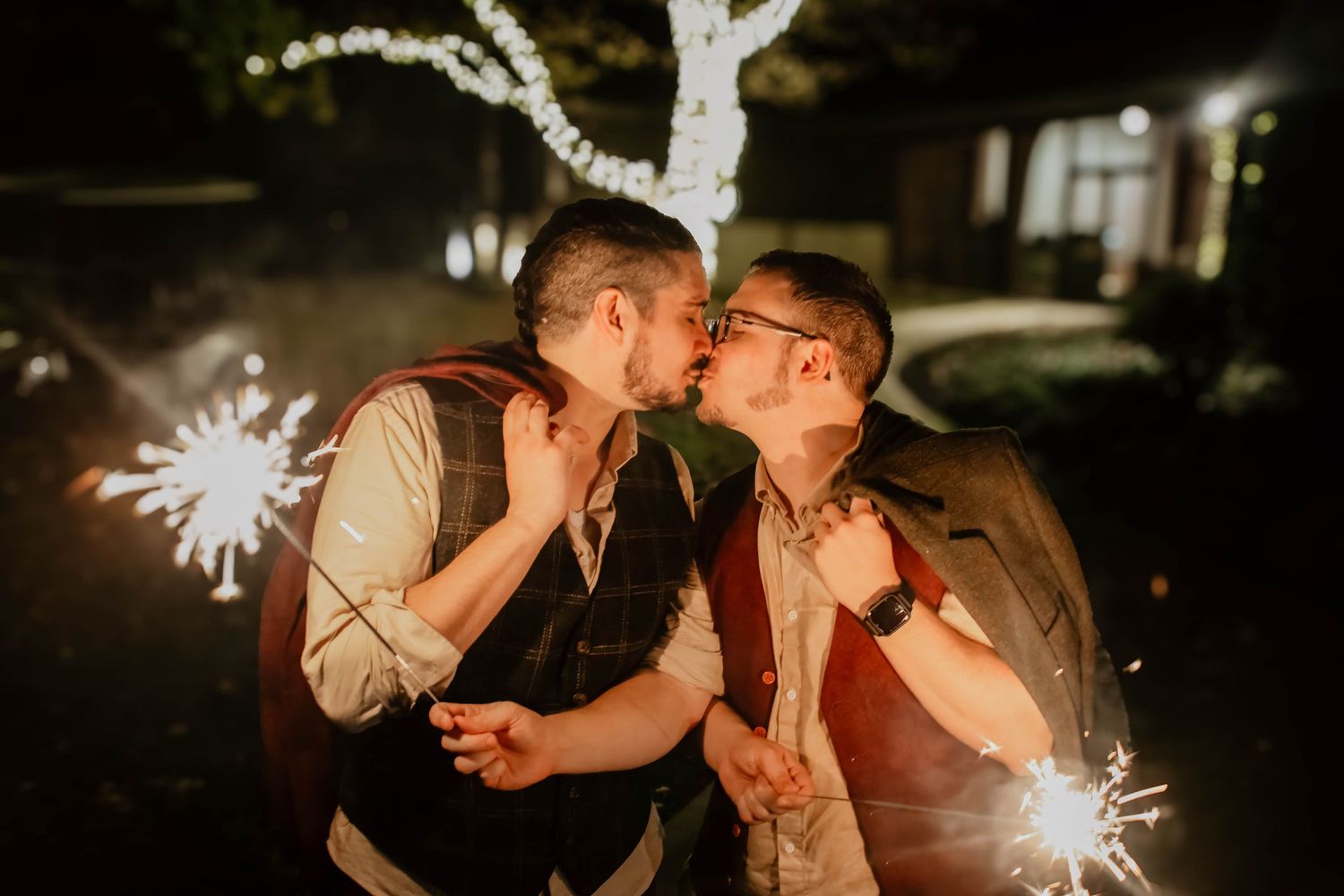 A couple shares a romantic kiss while holding sparklers in front of illuminated trees at night.