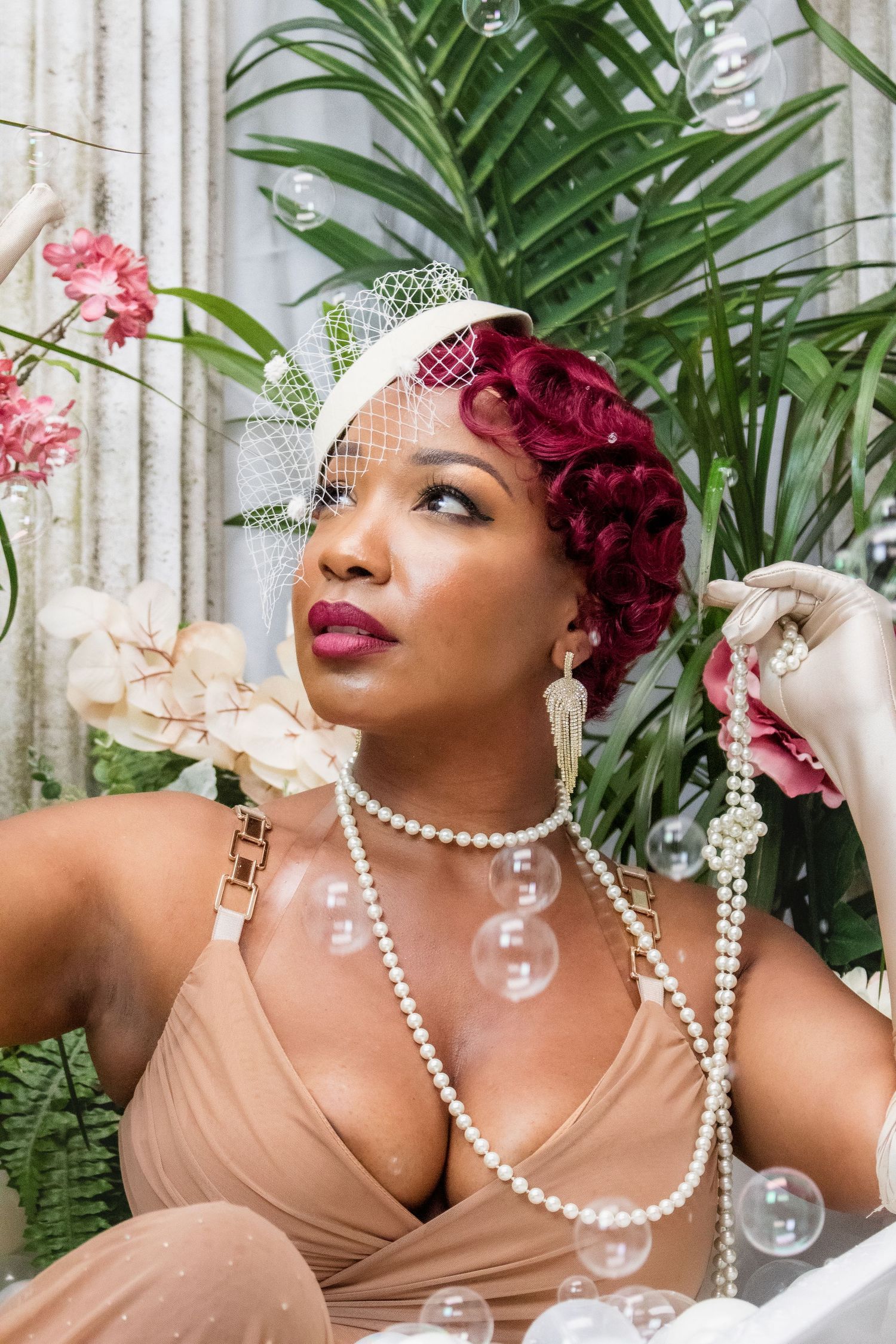 A glamorous vintage-inspired portrait with pearls, burgundy flowers, and tropical plants against a white backdrop.
