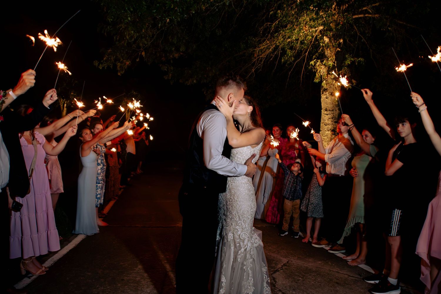 Couple embraces for romantic sparkler exit during nighttime wedding celebration as guests hold sparklers creating magical tunnel.