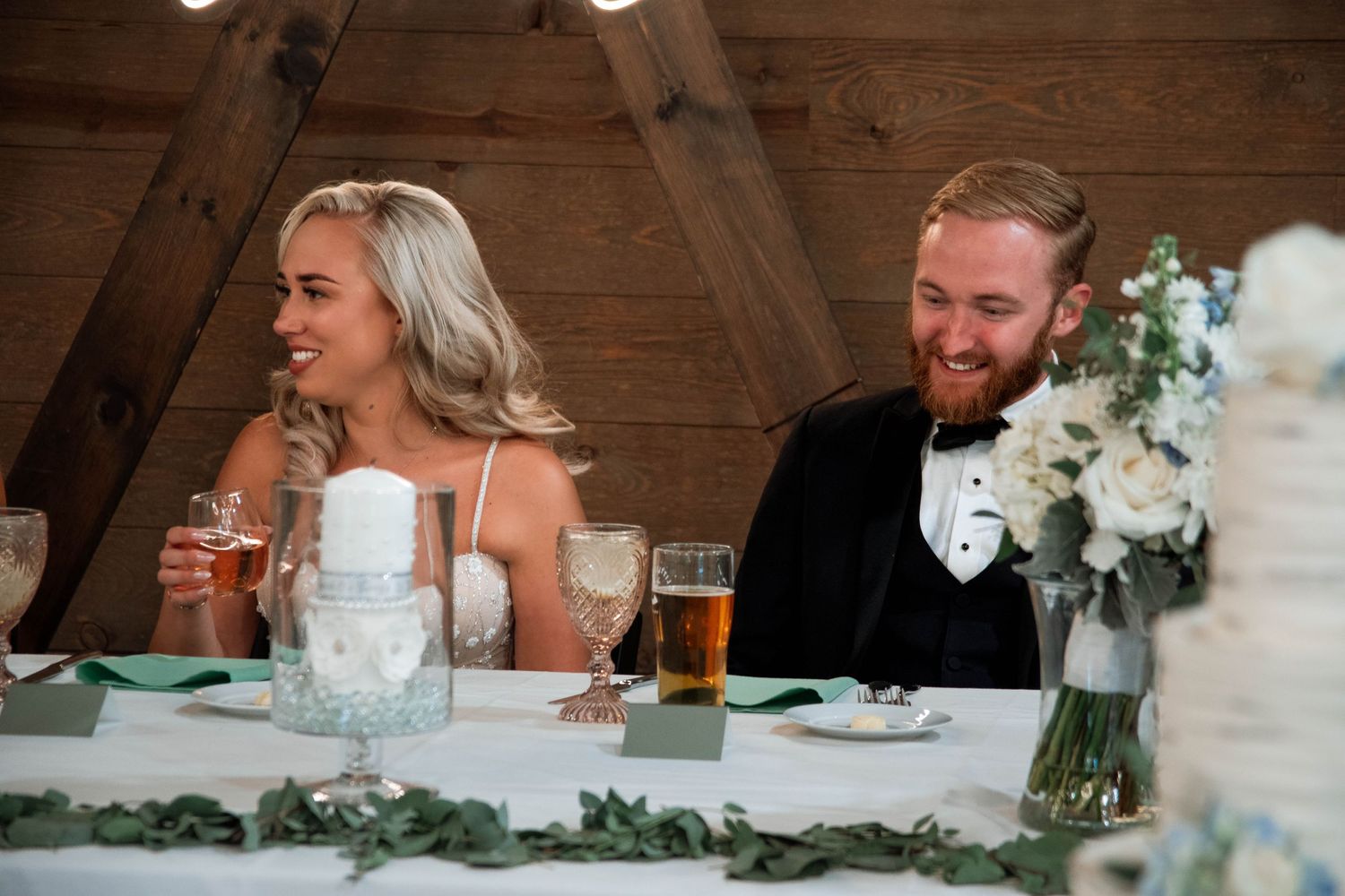 A couple shares a laugh during their wedding reception at a rustic barn venue with white floral decorations.