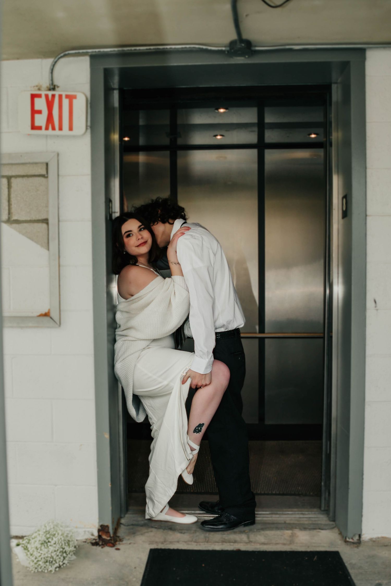 A romantic elevator moment with couple in wedding attire, with dramatic lighting and an exit sign above.