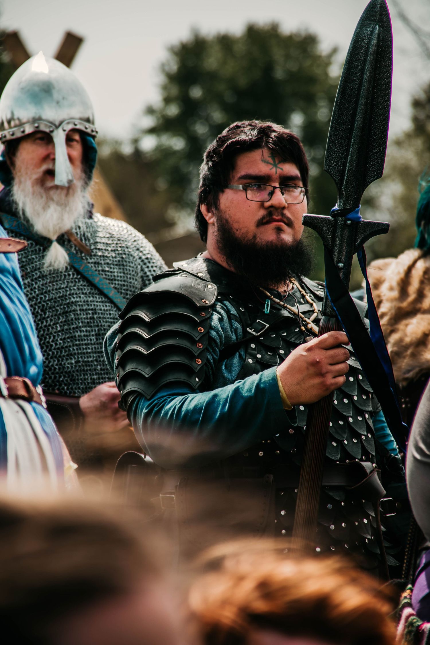 Medieval-style warrior wearing chainmail armor and glasses stands at attention with sword outdoors.