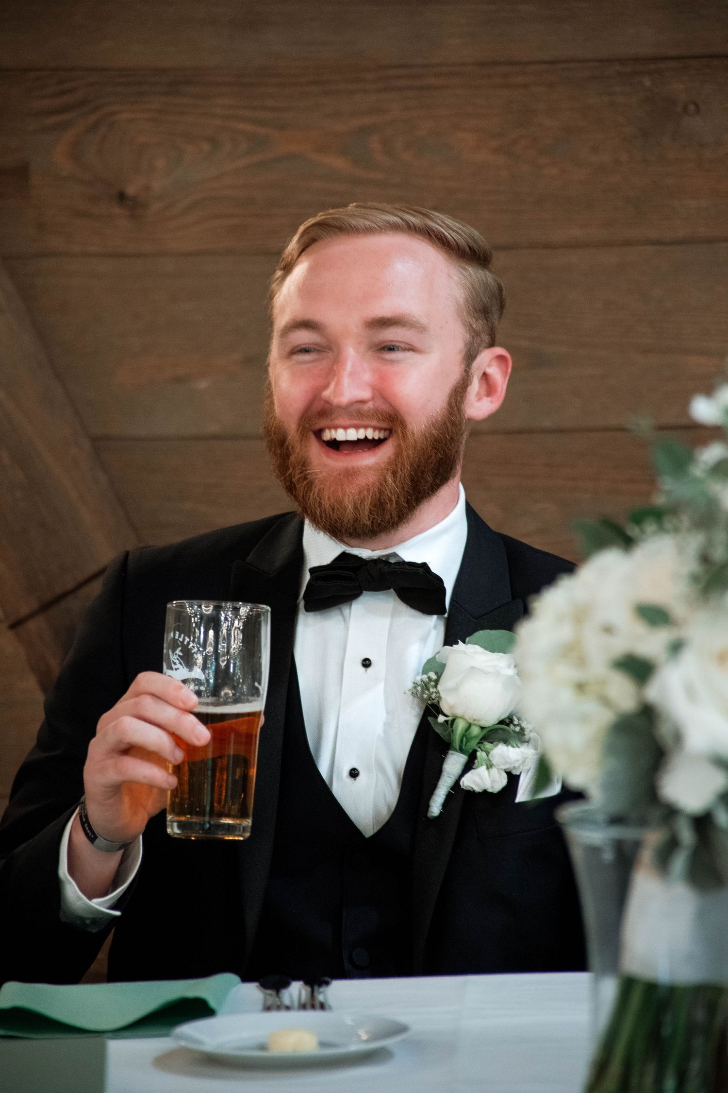 A groom in a black tuxedo laughs while holding a beer at his wedding reception next to white floral centerpieces.