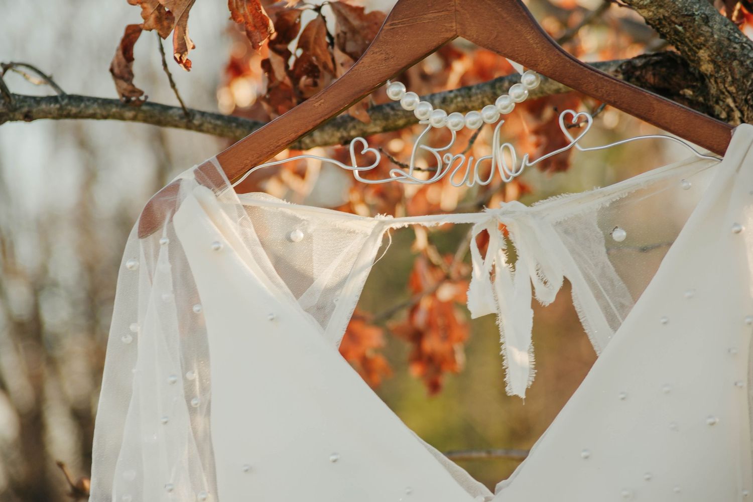 White lace wedding dress hangs on wooden hanger against autumn foliage backdrop during golden hour.