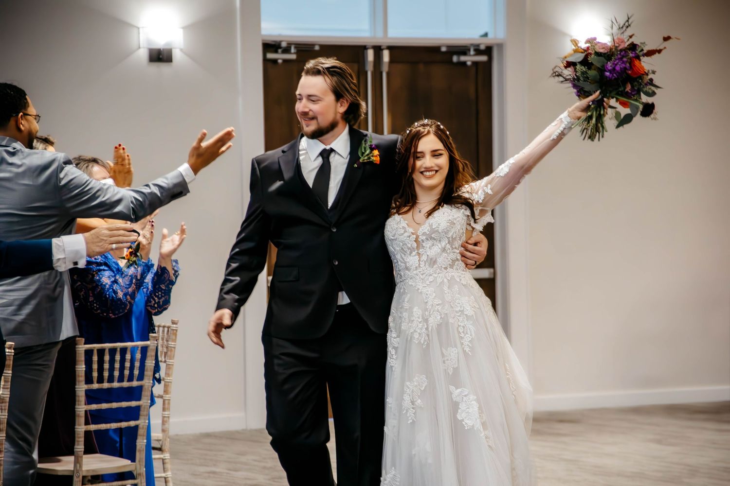 A joyful couple in formal wedding attire celebrate as they walk down the aisle while guests cheer.