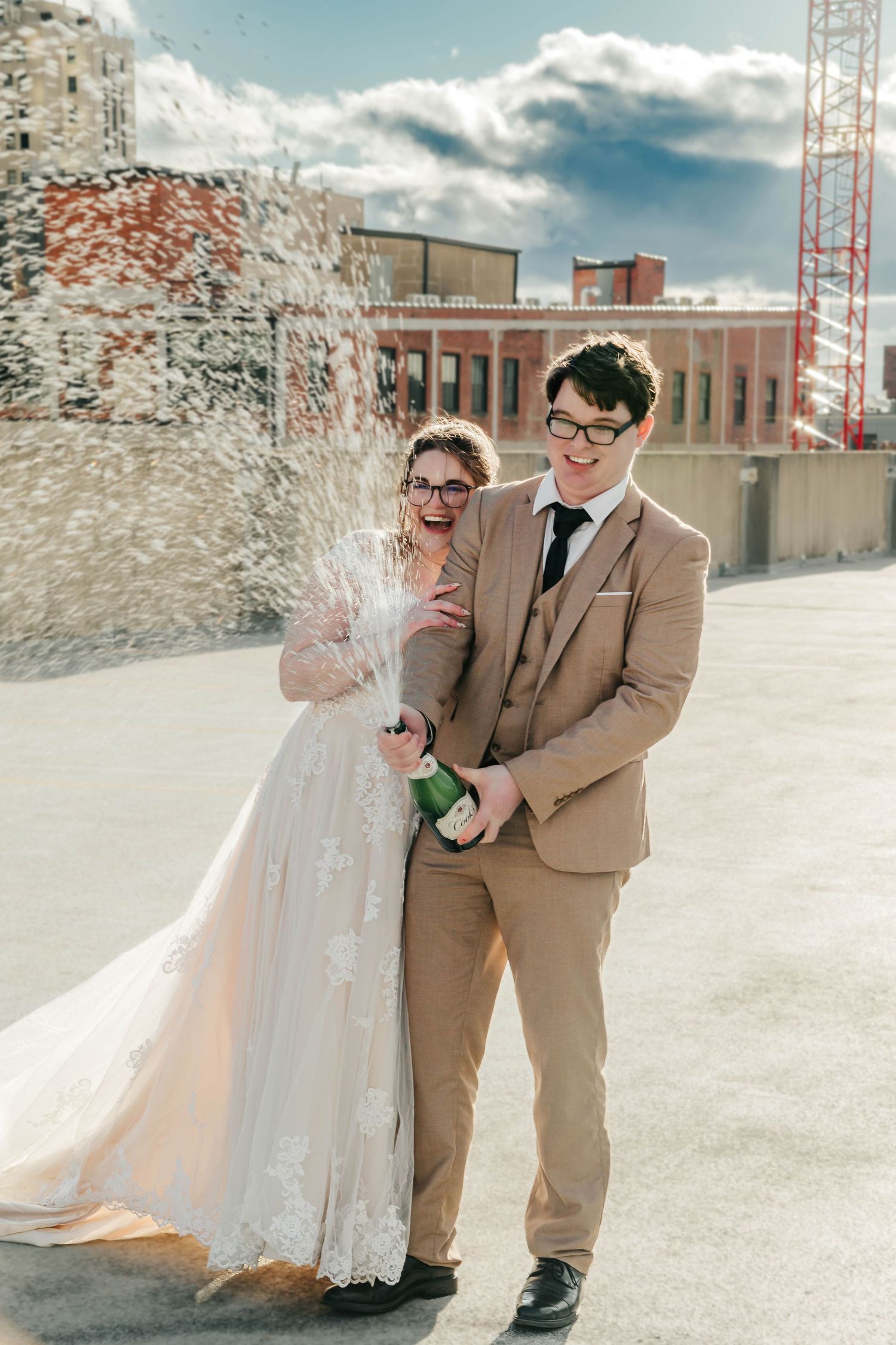 Newlyweds celebrate with champagne spray on a rooftop with urban buildings and blue sky in the background.