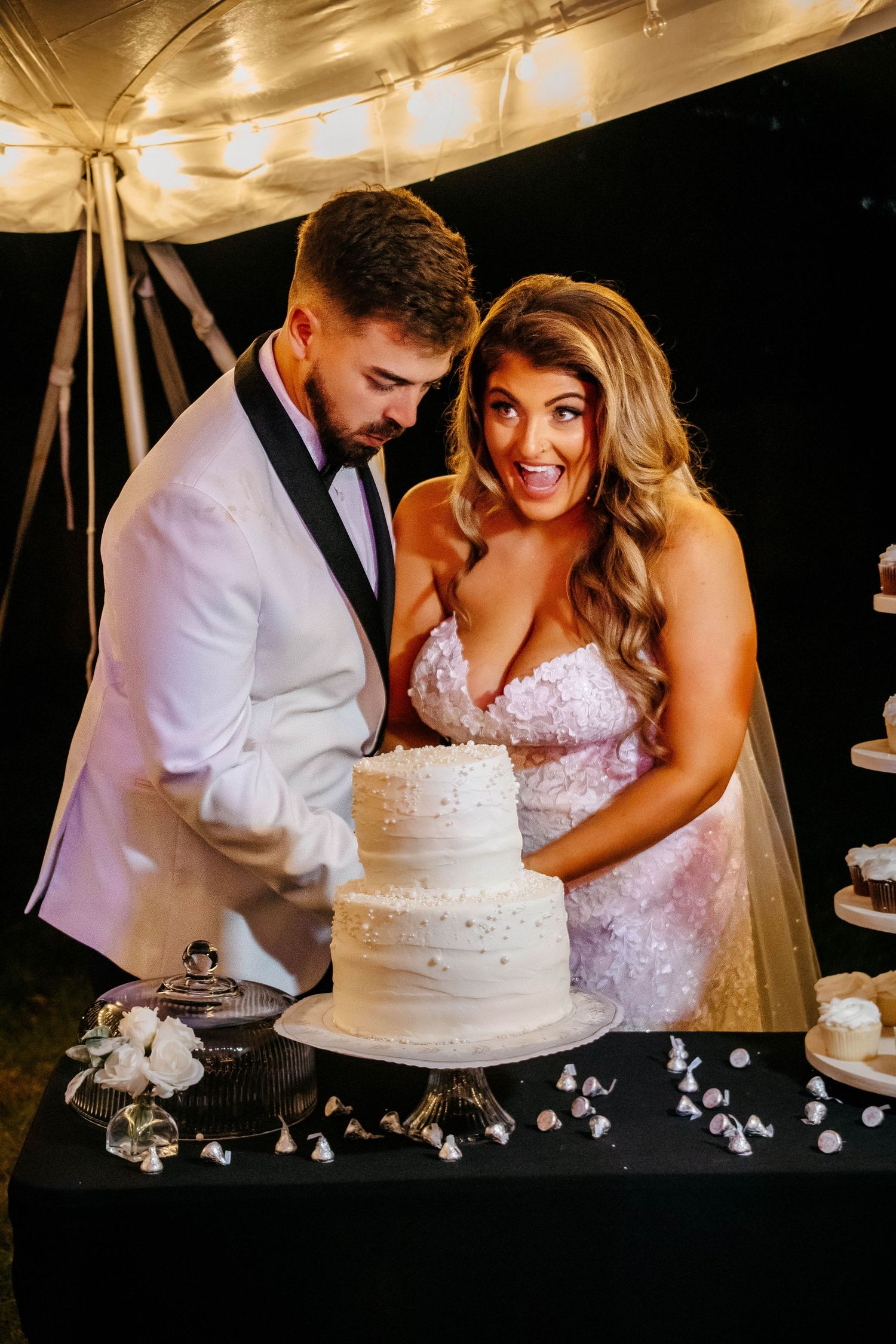 A couple cuts their elegant white two-tier wedding cake under a tent with warm lighting.