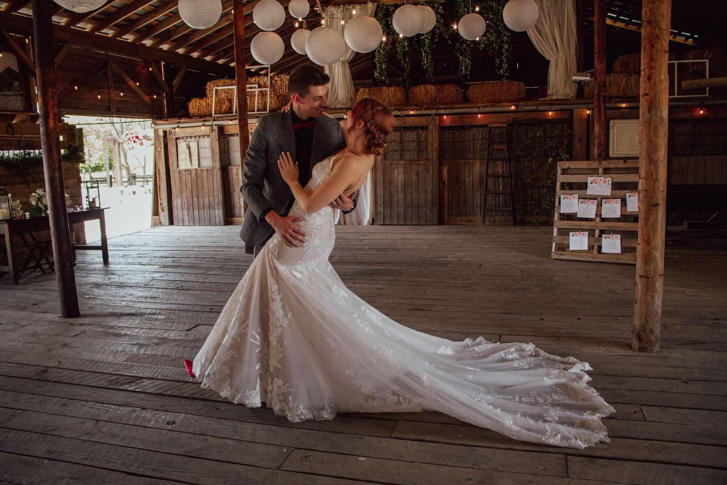 A couple shares an intimate moment dancing on a rustic wooden barn floor decorated with hanging white lanterns.