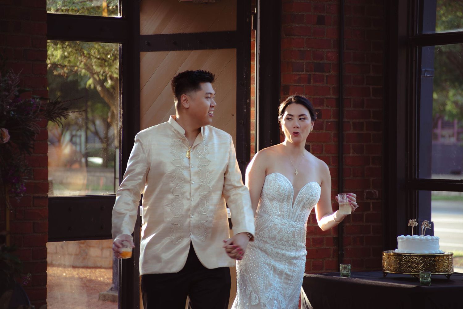 A couple in formal attire stands together at a rustic brick venue with natural lighting streaming through windows.