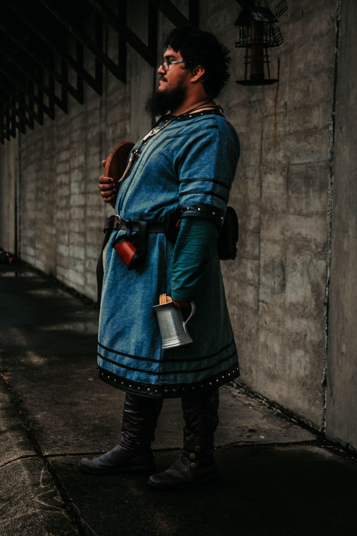 Figure in a blue denim coat and black boots stands contemplatively against a concrete wall in moody lighting.