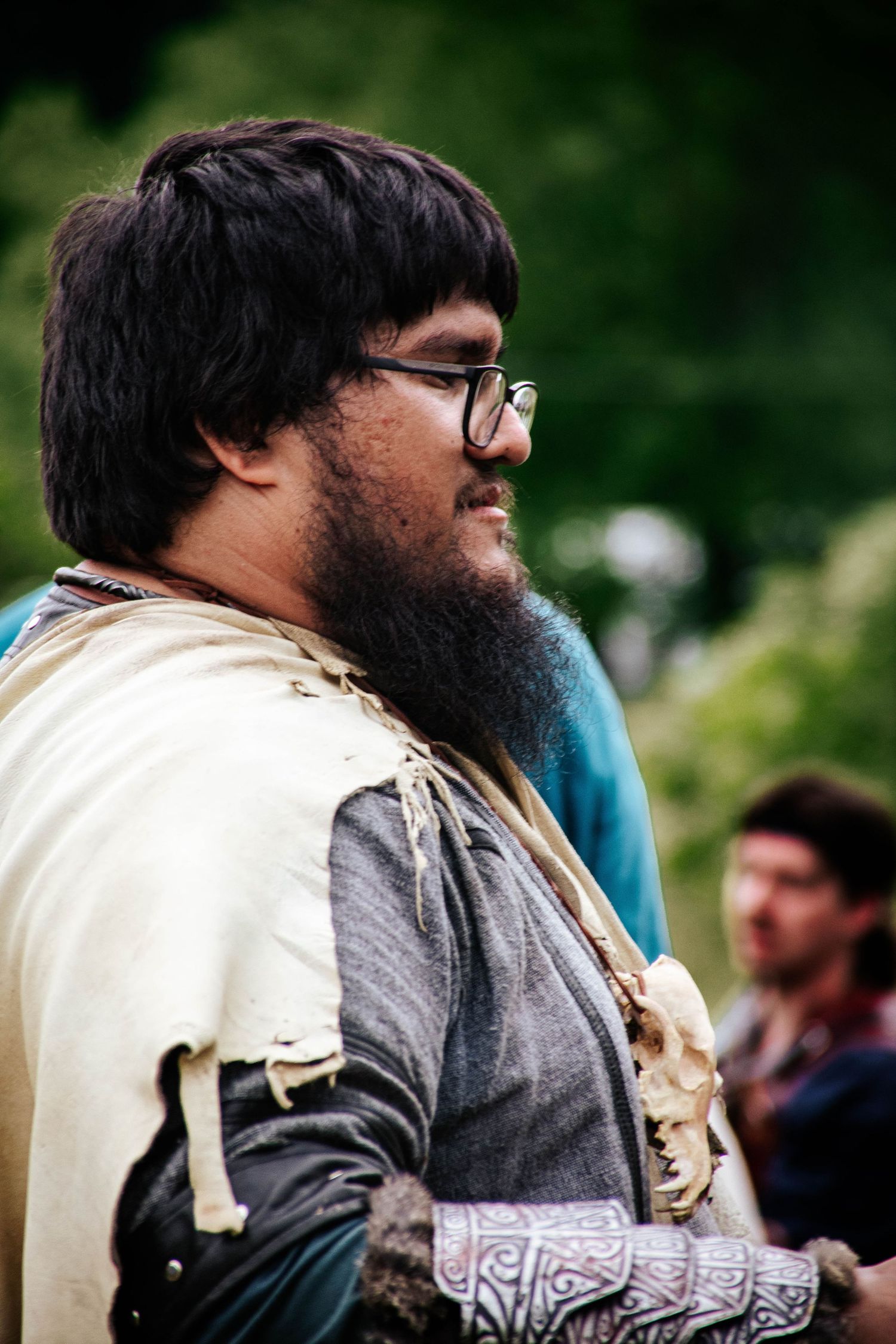 A person wearing glasses and a thick beard with a beige vest is shown in close-up profile view at an outdoor event.