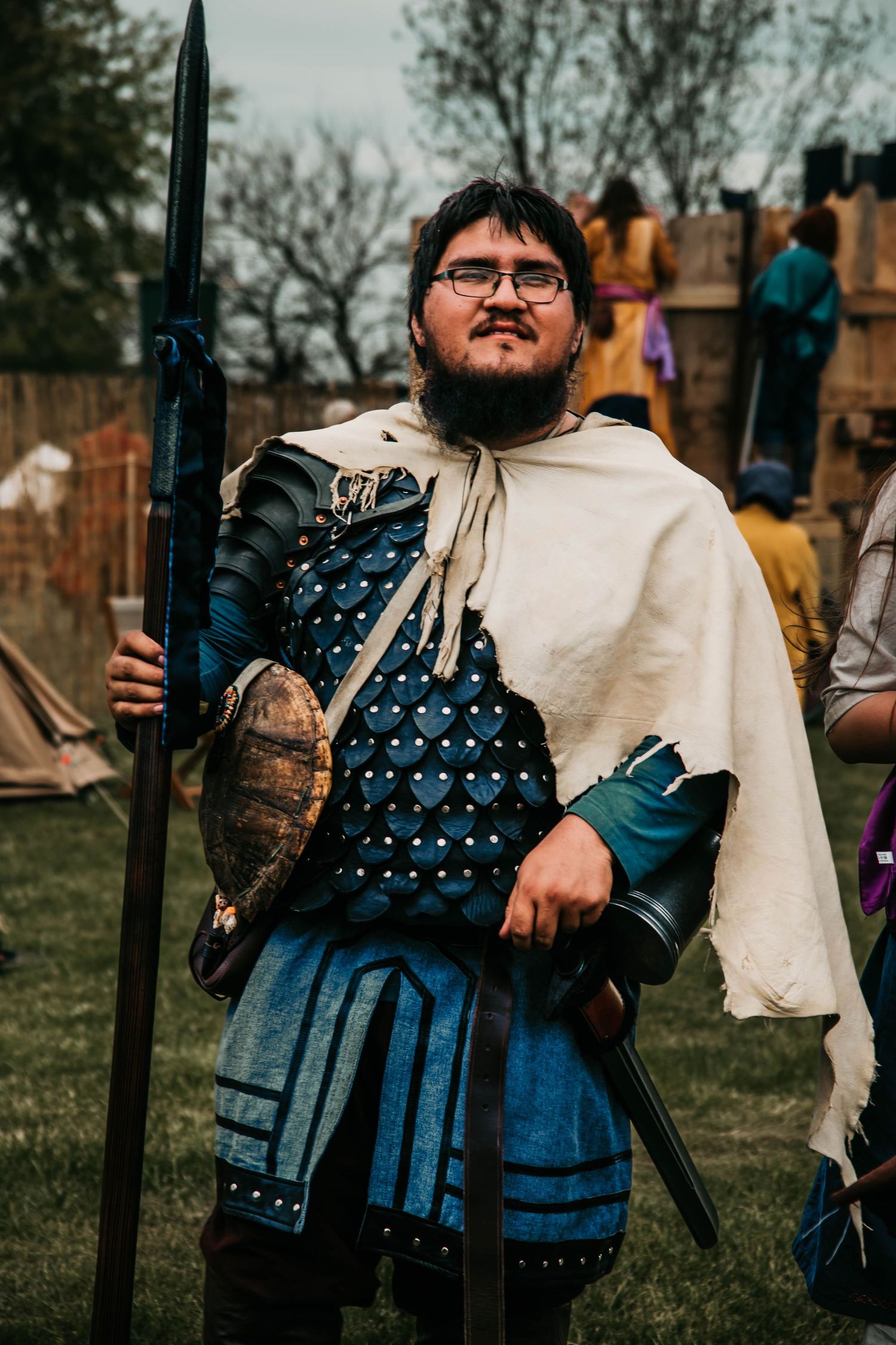 Medieval reenactor in studded blue leather armor and white cloak holds spear at renaissance faire.