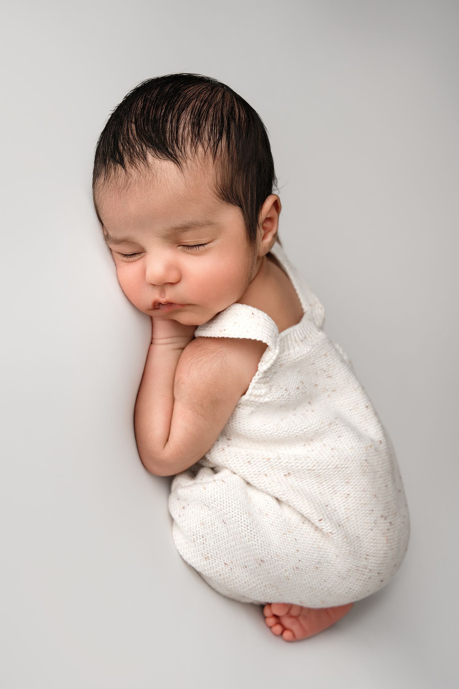 A sleeping newborn baby curled up in white clothing against a light gray background.