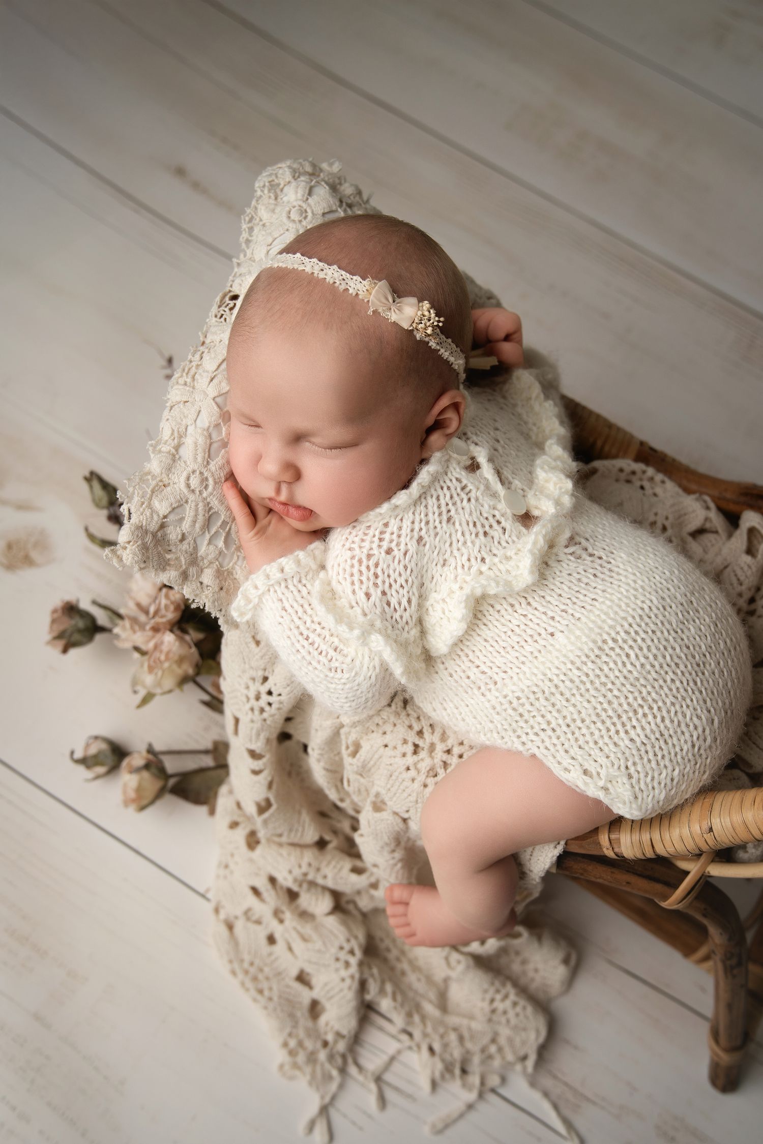 Sleeping newborn wearing white knit romper rests on vintage wooden surface with lace and floral accents.