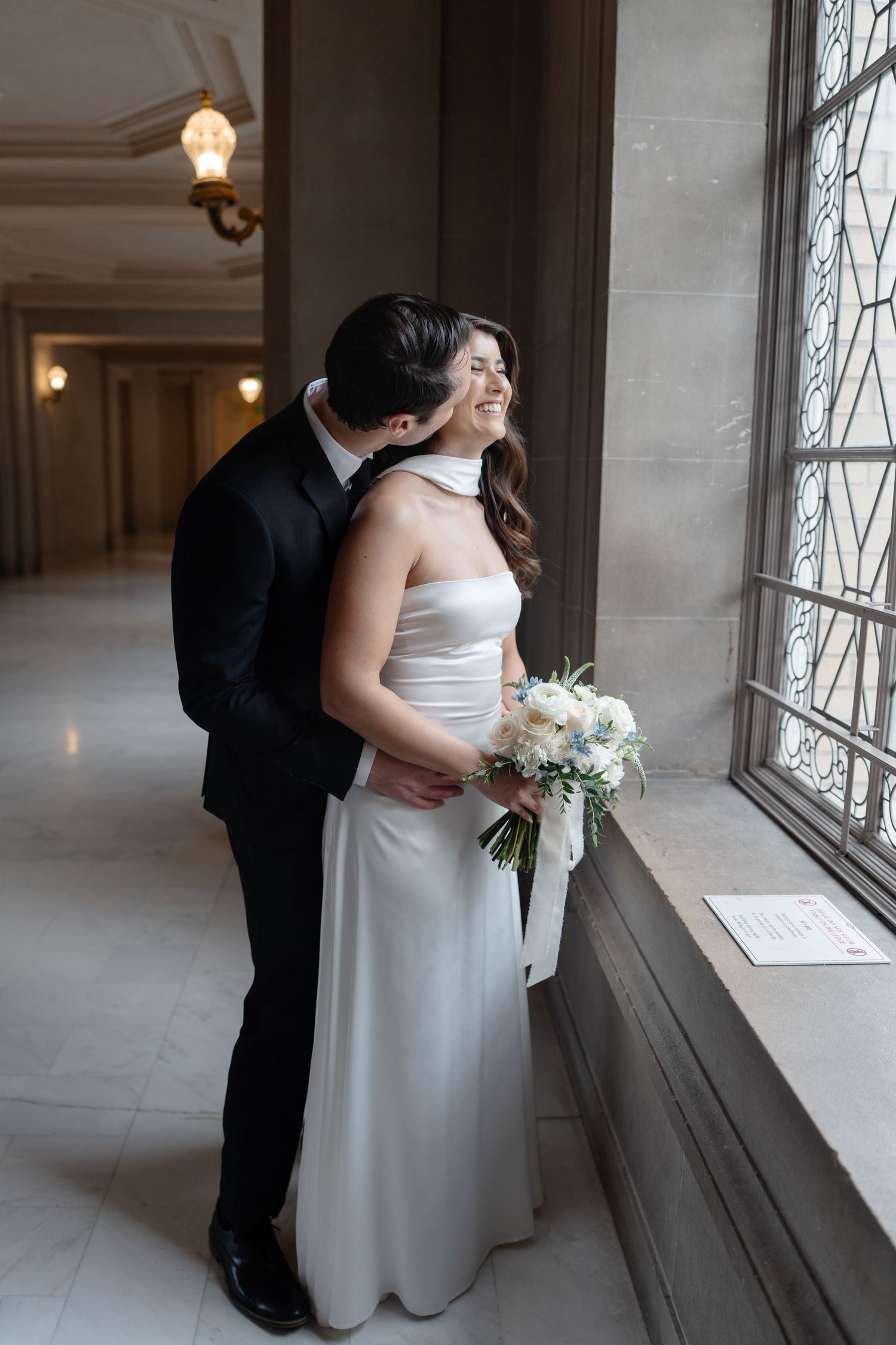 Elegant couple shares intimate moment by ornate window in classic venue with white wedding gown and bouquet.